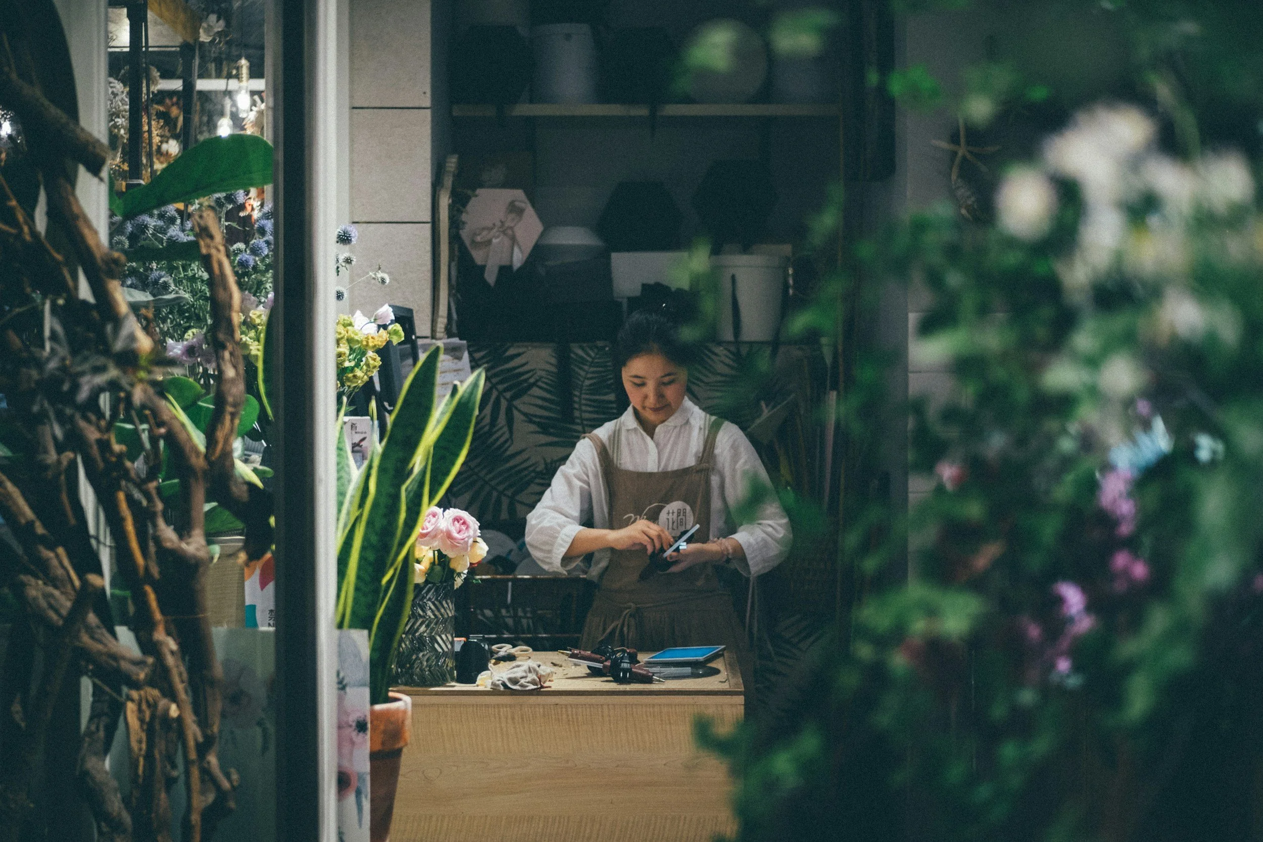 An SME business owner who secured a loan in a white shirt and apron is working behind a counter in a flower shop, surrounded by various plants and flowers, as seen through a window.
