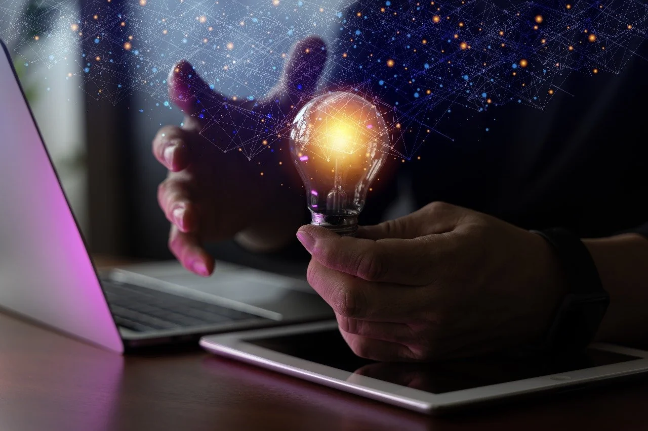Person holding a glowing lightbulb with a digital network overlay, sitting at a desk with a laptop and tablet.