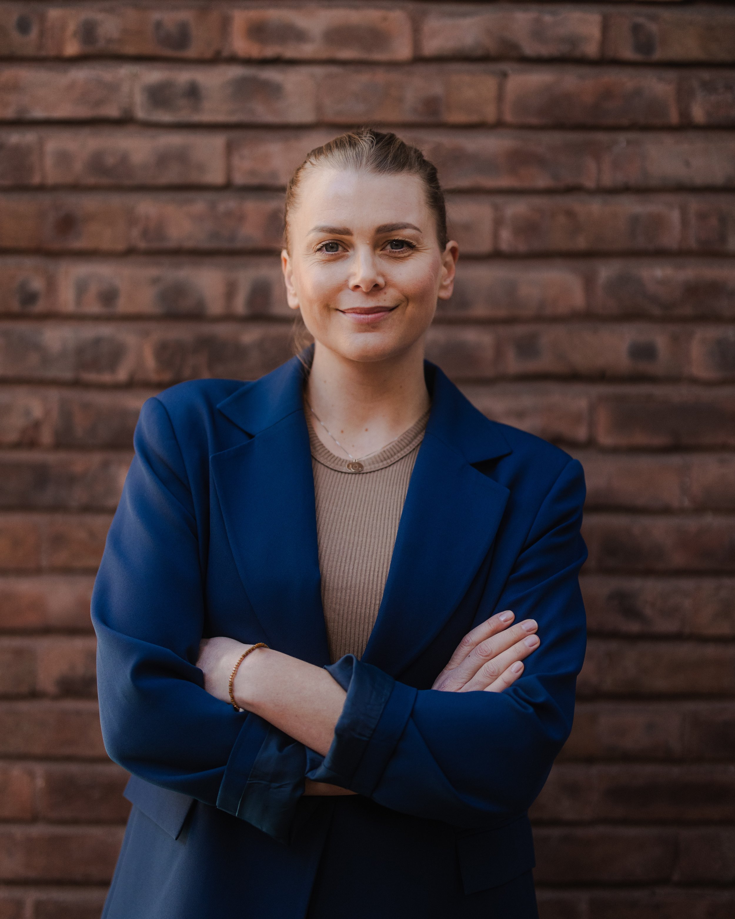 Kvinna med korsade armar, ljus hud, ljusbrunt hår i hästsvans, klädd i blå kavaj och beige topp, står framför en tegelvägg.