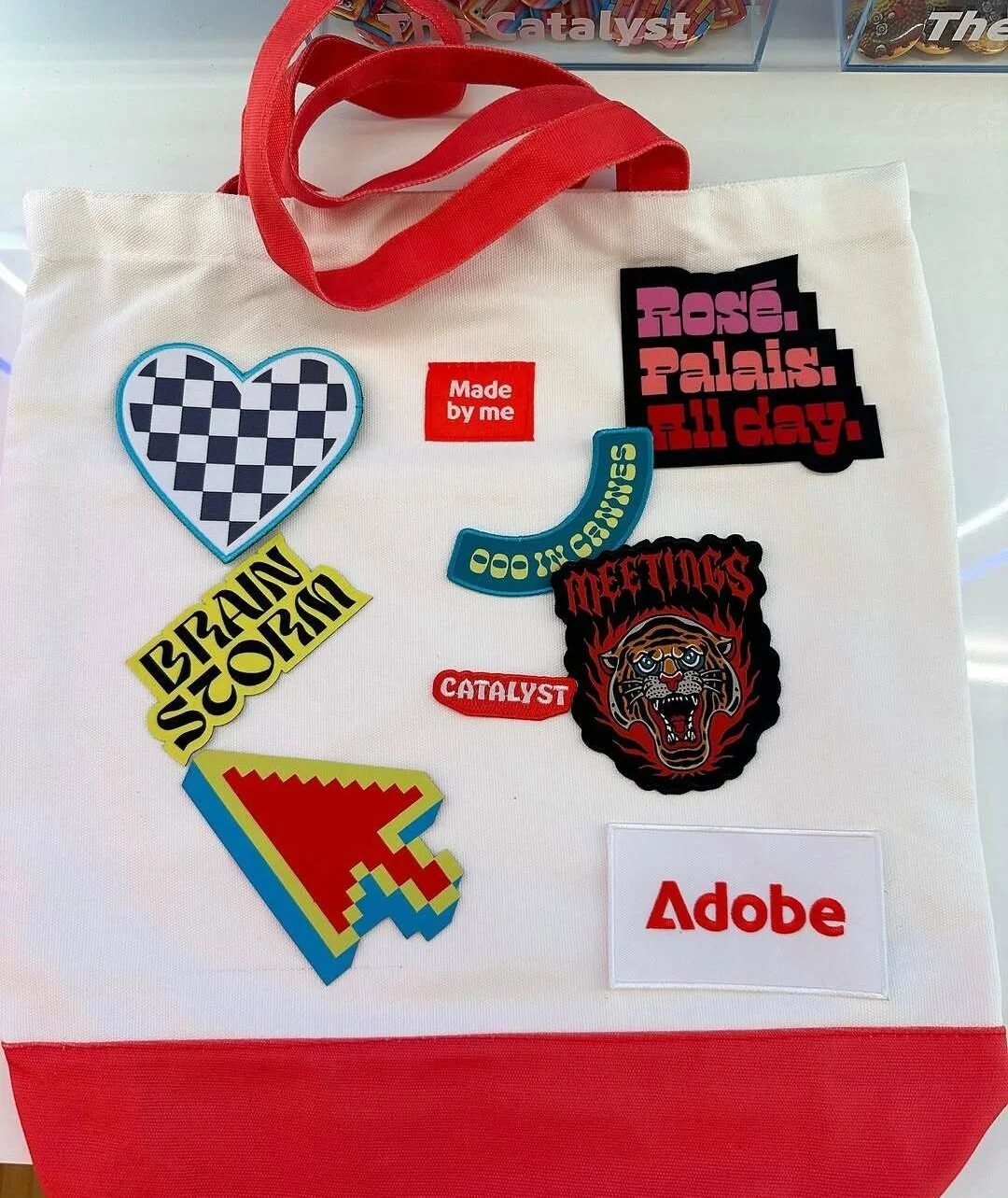 A white tote bag with red handles decorated with various colorful patches and stickers, including a checkered heart, a yellow patch with black text, a pixelated arrow, and patches with phrases like 'Made by me', 'Rose. Palais. I'll stay.', 'Let's go', 'Meetings', 'Catalyst', and 'Adobe', along with a tiger face patch.