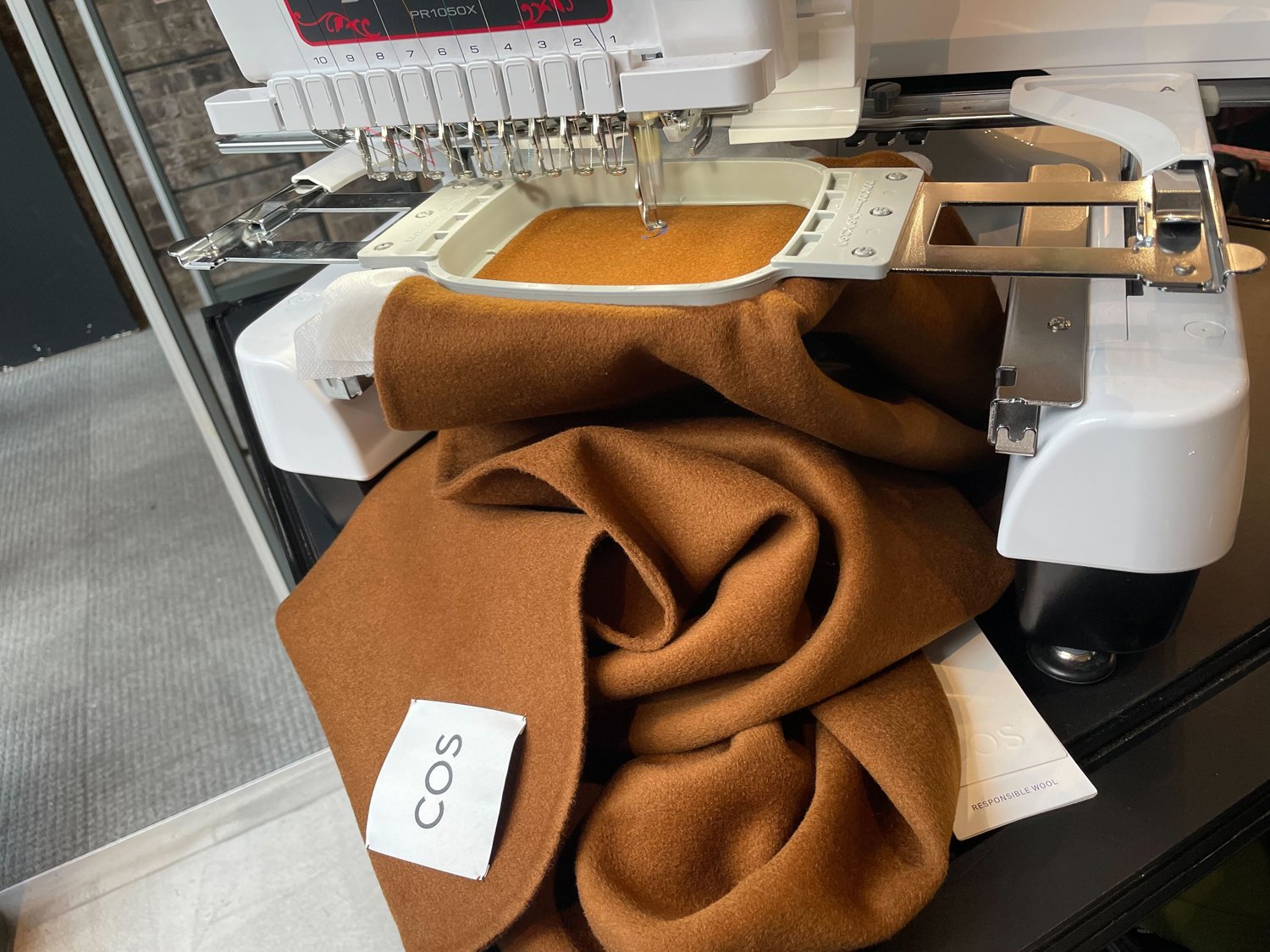 A wool blanket is being embroidered with a sewing machine, with a white tag labeled "COS" attached to it.