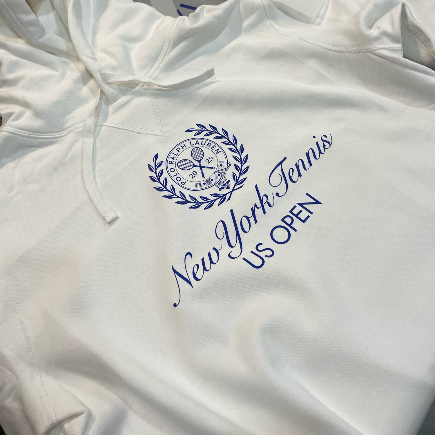 White hoodie with printed emblem and text for the New York Tennis US Open, featuring a tennis ball, racquets, and laurel wreath design.