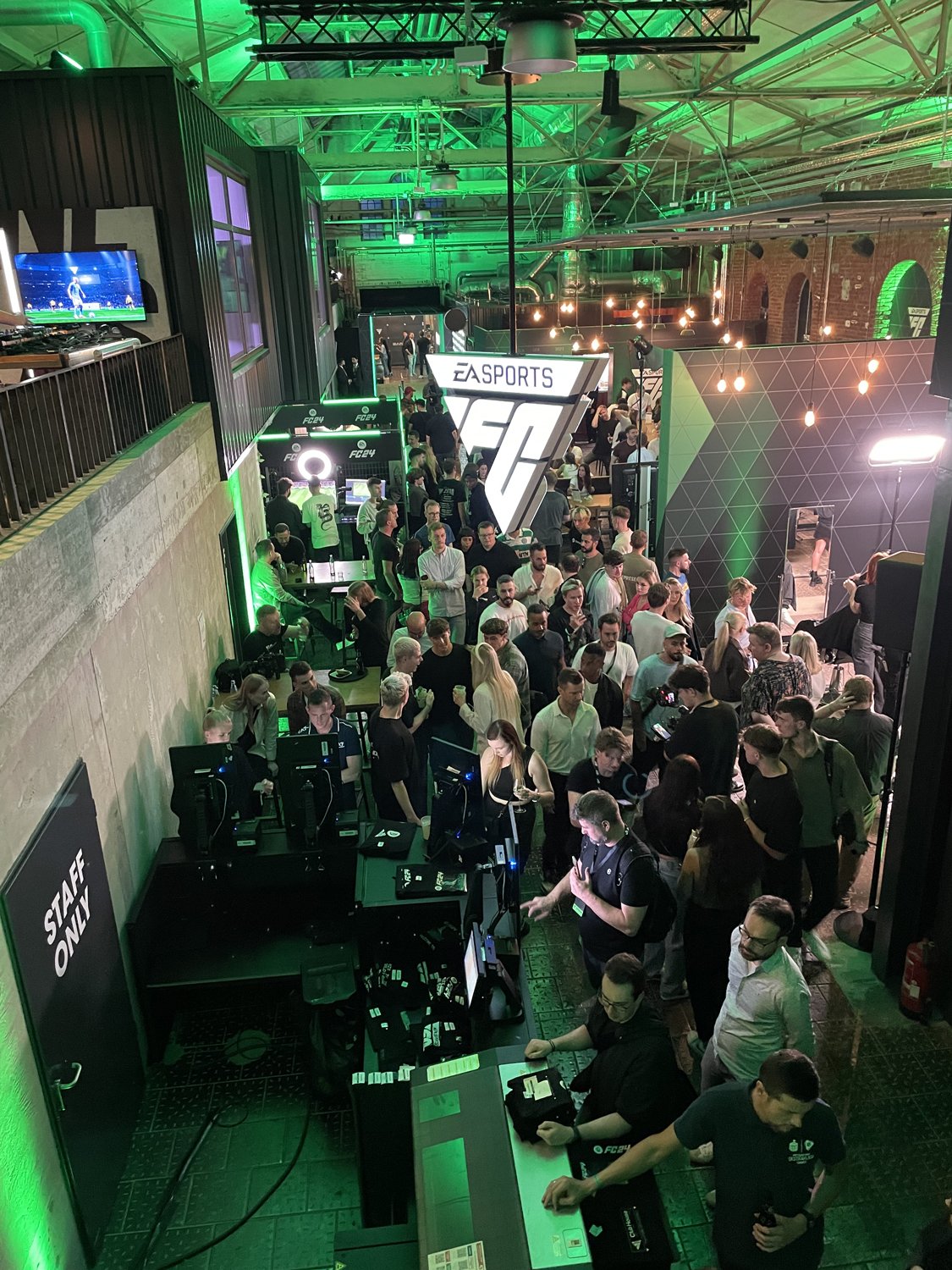 A busy indoor event with many people, gaming stations, and EA Sports FC related signage. Green lighting and neon signs create a vibrant atmosphere.