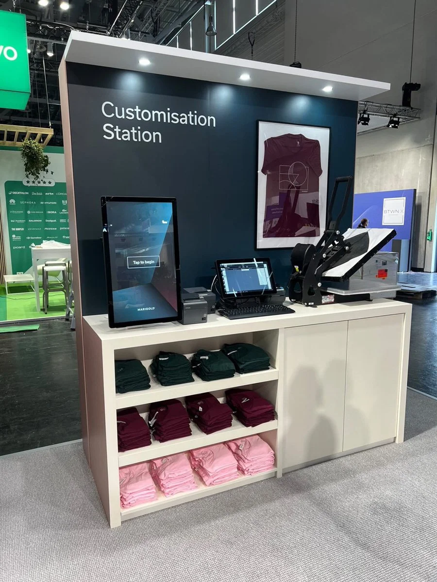 A customization station at a retail event with a touchscreen, printer, and computer setup, shelves with folded t-shirts in black, maroon, and pink, and a framed graphic of a t-shirt on the wall.