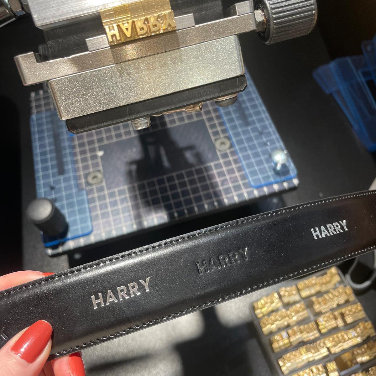Close-up of a black strip with the word "HARRY" repeated three times in silver, held by a person with red painted nails. In the background, a jewelry-making or watchmaking workspace with a metal tool, a cutting mat, and small compartments with gold items are visible.