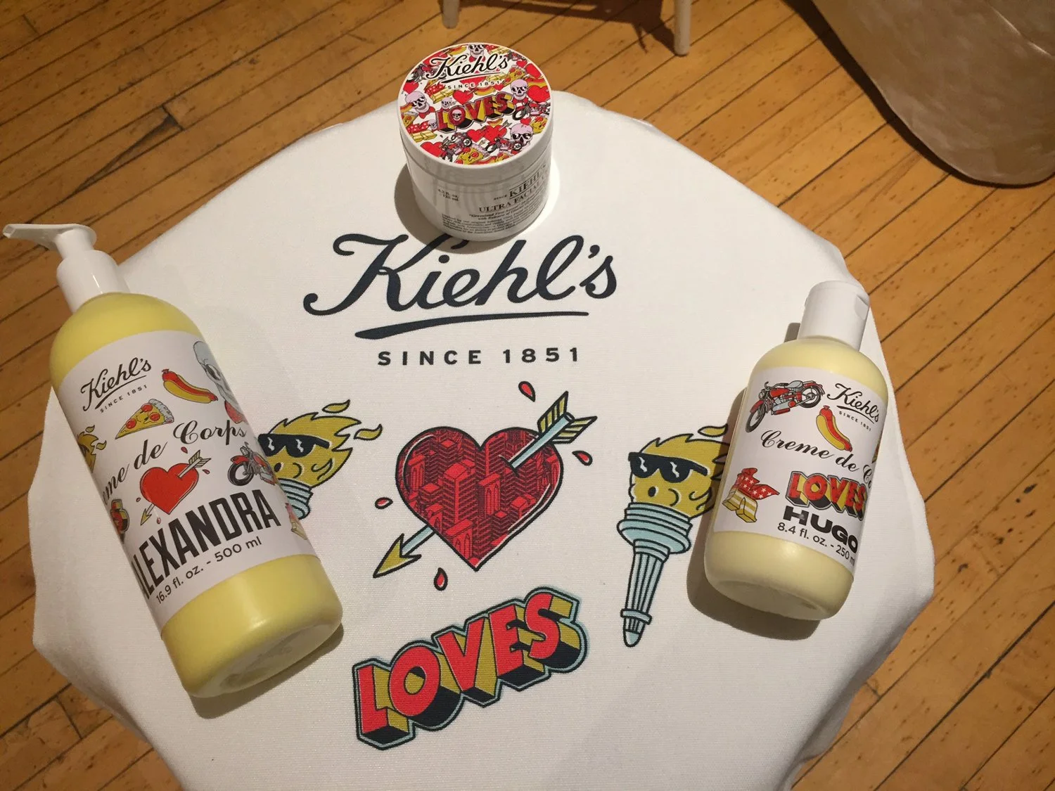 A Kiehl's branded cloth with Retro Love design, two bottles of Kiehl's creamy de leche lotions, and a small container of Kiehl's Love Loves label on a wooden floor.