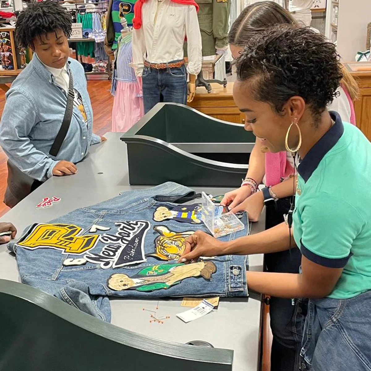 A woman is signing a denim jacket with patches and embroidery at a store counter, while a young girl watches. Other shoppers and clothing displays are in the background.