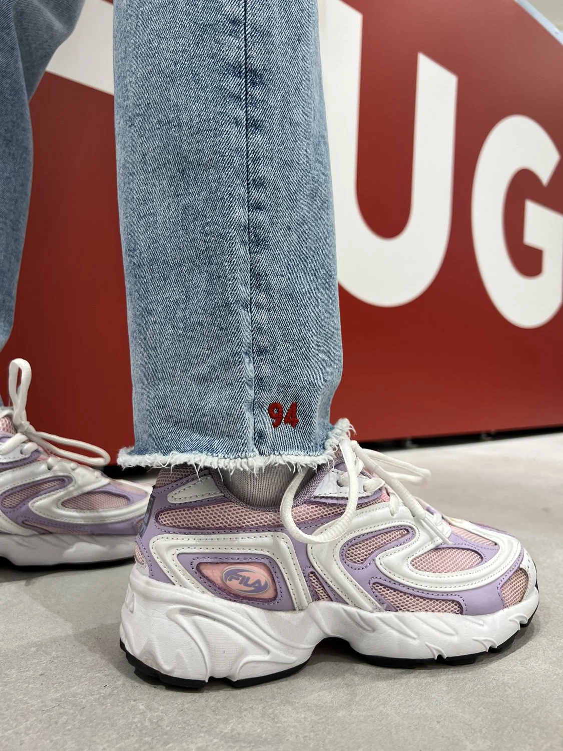 Close-up of a person's foot in a purple and white FILA sneaker, wearing light blue jeans with a frayed hem and red embroidered number '94' at the ankle, standing on a gray floor in front of a red background.