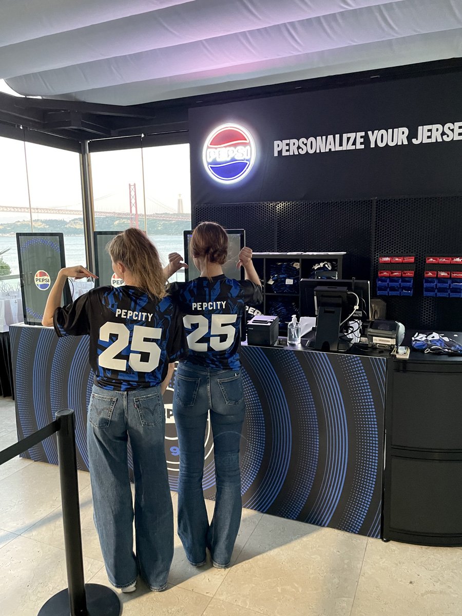 Two women wearing Pepecity jerseys with the number 25, standing at a Pepsi promotional booth near the water with a bridge visible in the background.