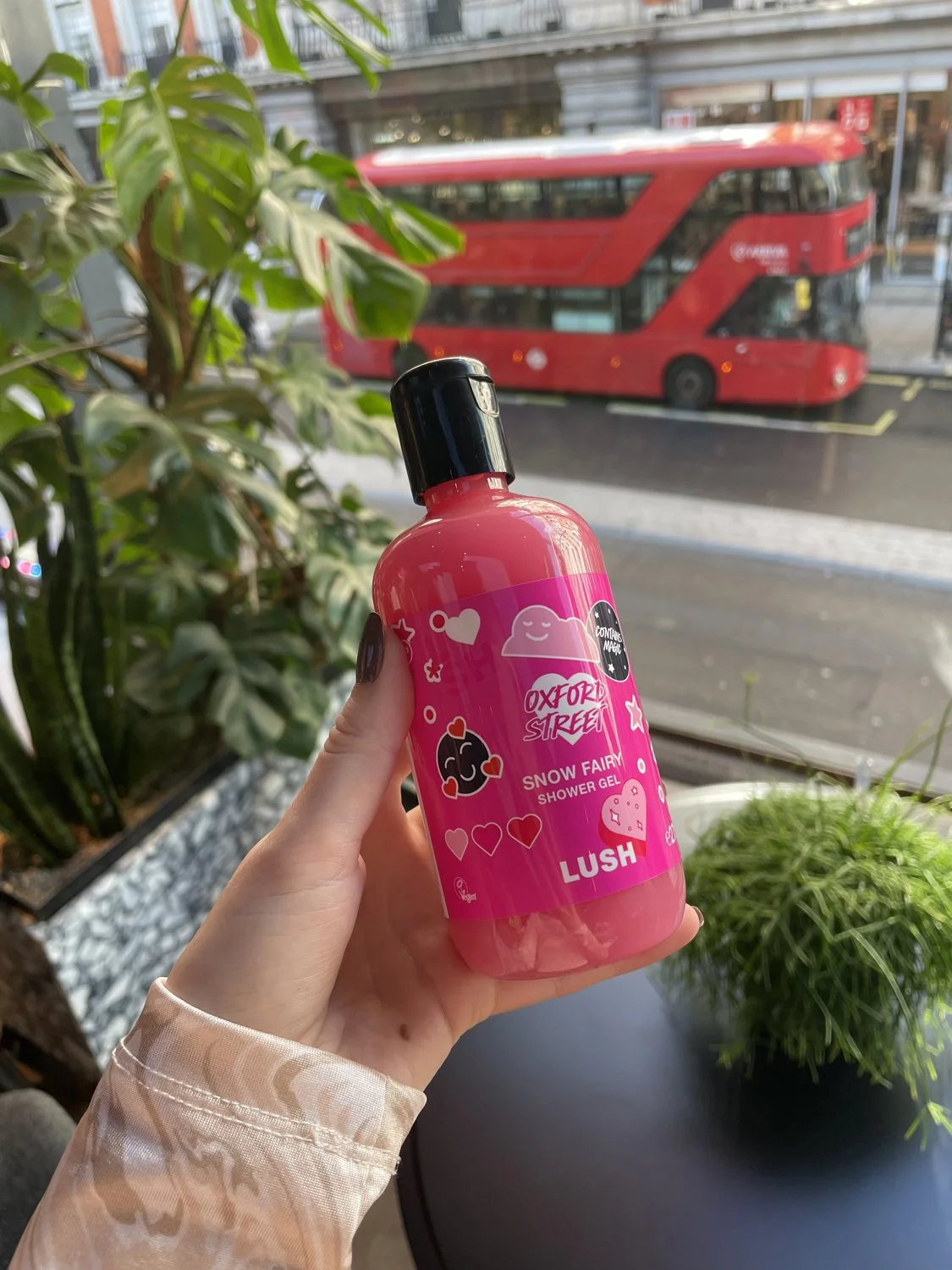 Pink bottle of Lush Snow Fairy shower gel labeled Oxford Street, held by a person with dark nail polish, with a city street and a red double-decker bus outside the window.