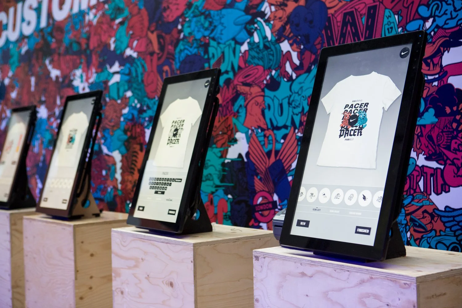 Multiple digital screens on wooden pedestals displaying T-shirt design customization with a graphic of a lion, eagle, and tiger, set against a colorful, graffiti-style mural background.