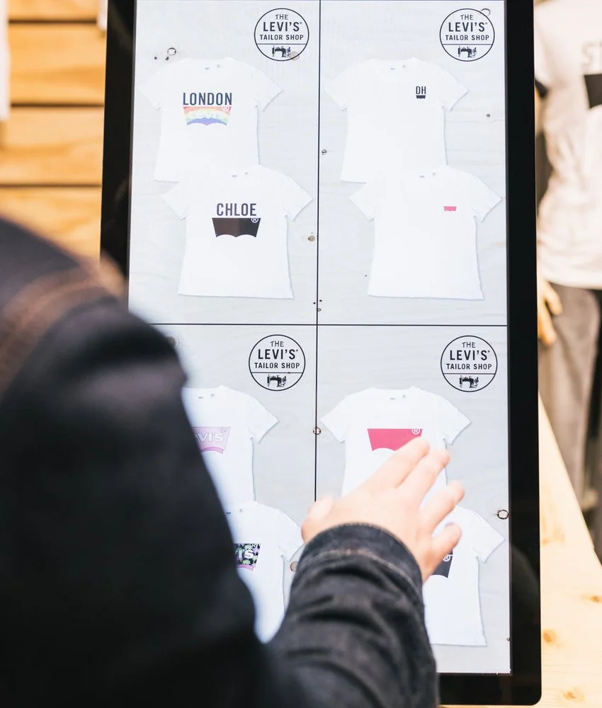Digital display showing various T-shirt designs for Levi's Tailor Shop, including designs labeled London, Chloe, DH, and one with pink and black graphics, with a person reaching out to touch the screen.