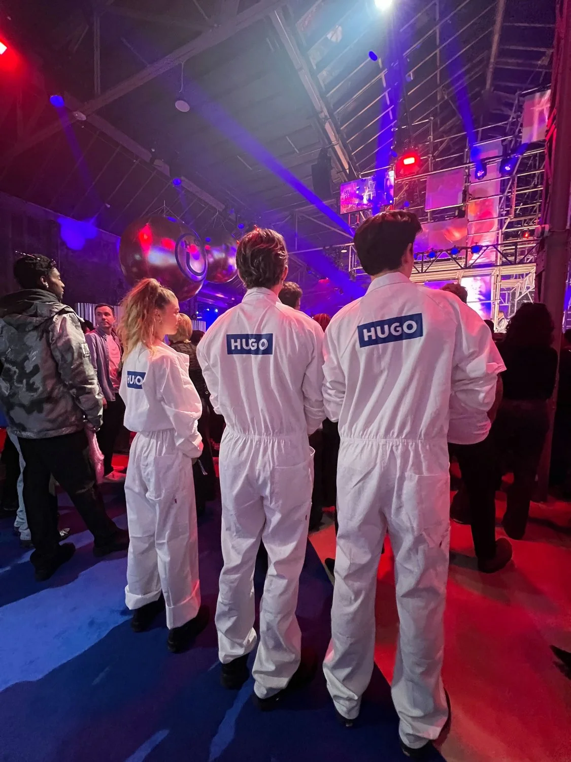 Four people wearing white jumpsuits with 'HUGO' labels on the back are standing in a dimly lit venue with colorful stage lights, large reflective balls, and a crowd of people watching a performance or event.