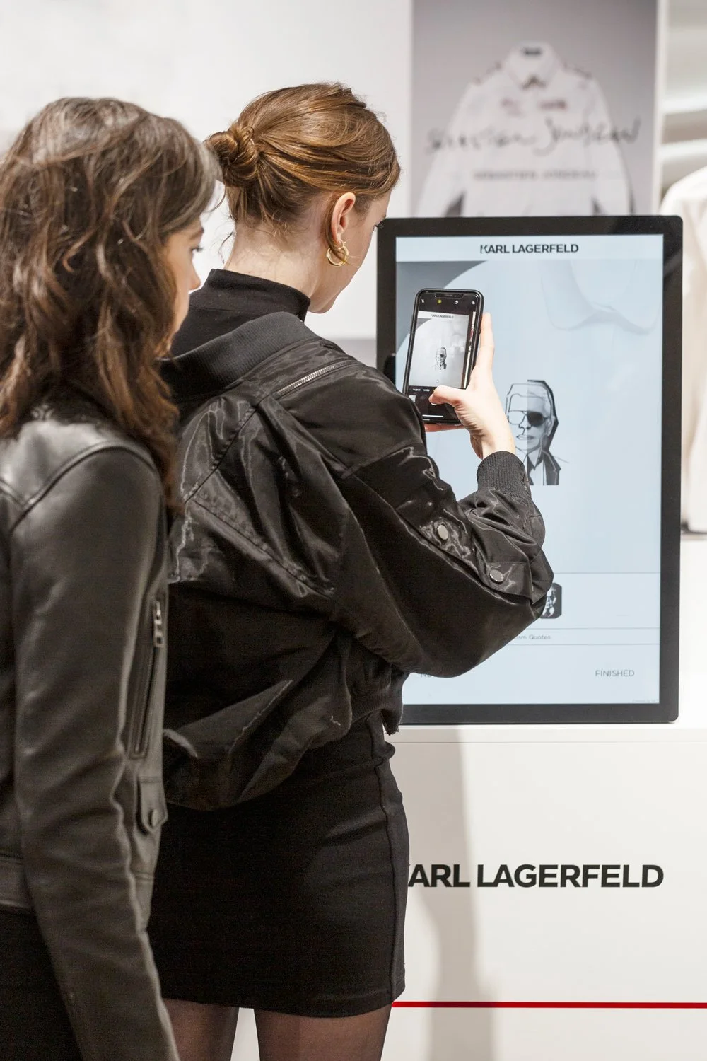 Two women standing in front of a screen displaying Karl Lagerfeld images, with one woman taking a photo of the screen with her phone.