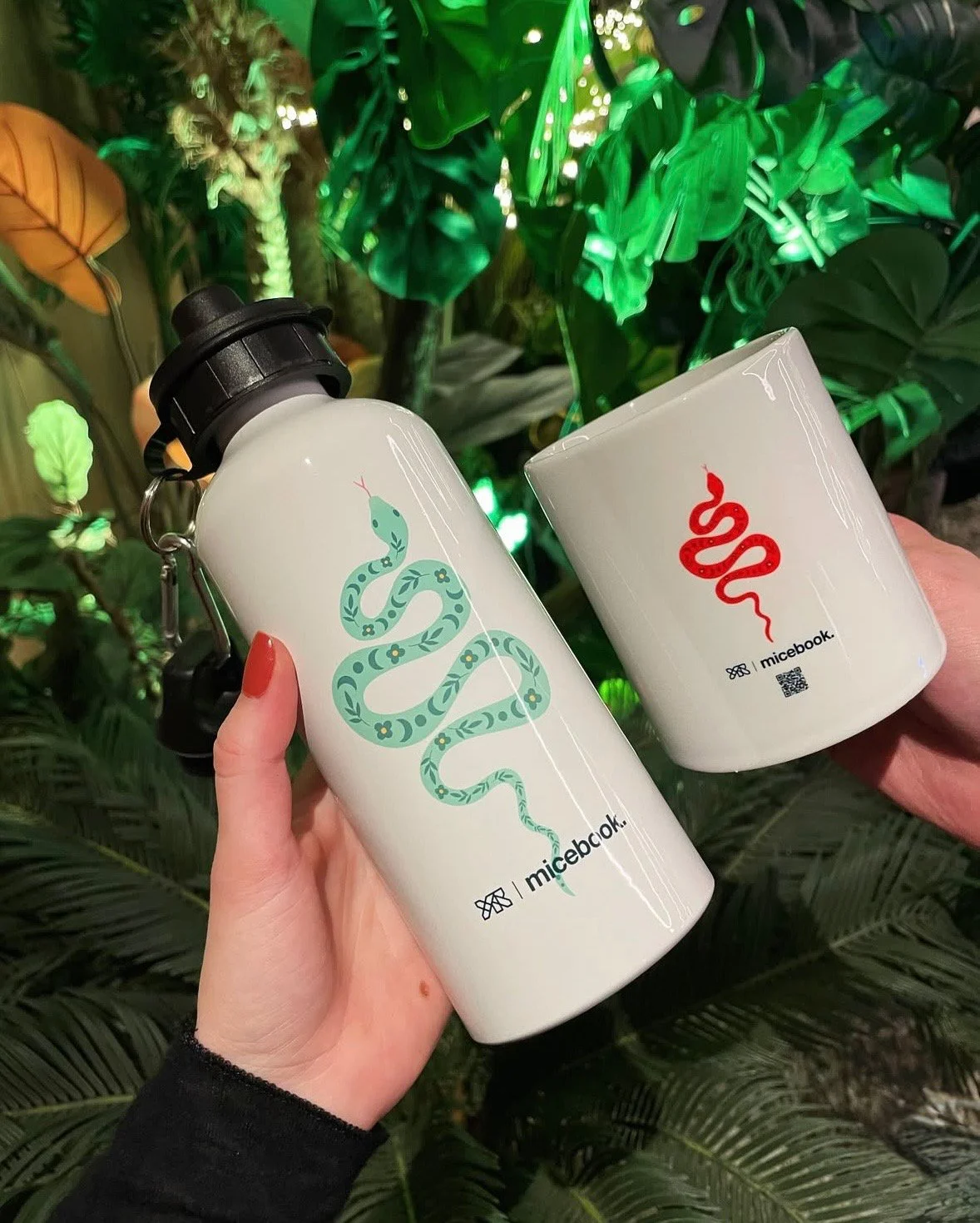 A person holding a white water bottle with a blue snake design and the word 'micebook' printed at the bottom, and another person holding a white mug with a red snake design and the same logo, with green tropical plants in the background.