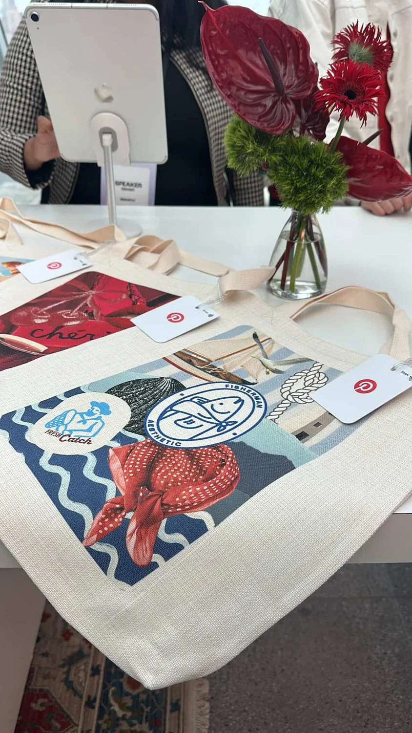 A table with a tote bag featuring colorful patches and logos, a vase of red flowers, and a person holding a tablet in the background.