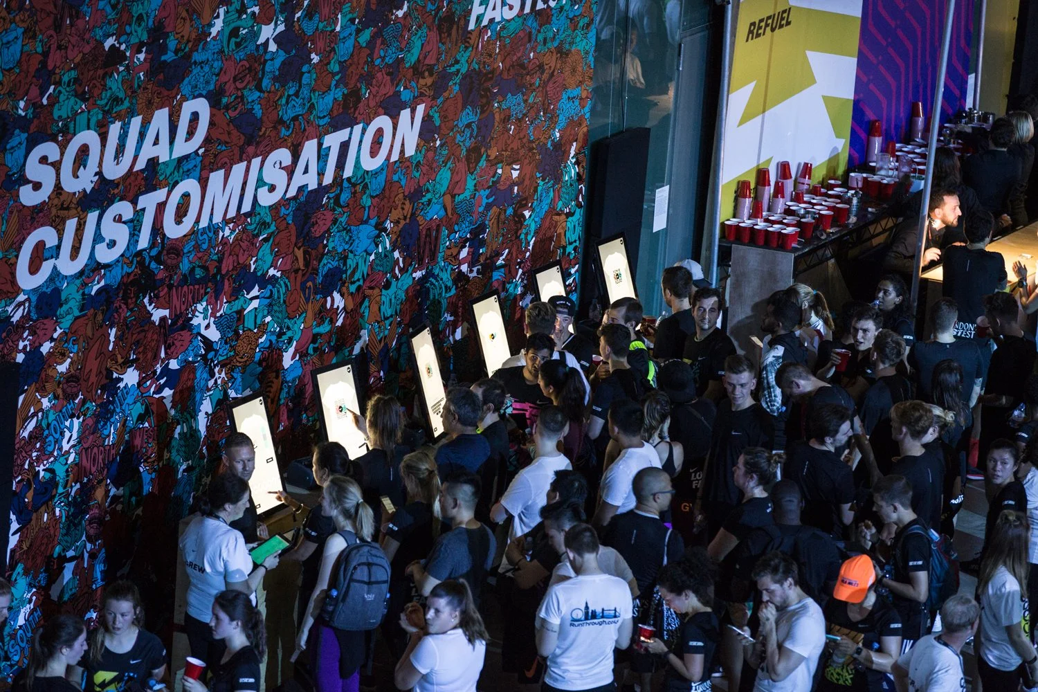 A large crowd of people waiting in line at a registration area with digital kiosks, a colorful mural with the words 'SQUAD CUSTOMISATION,' and a refreshment station with red cups.