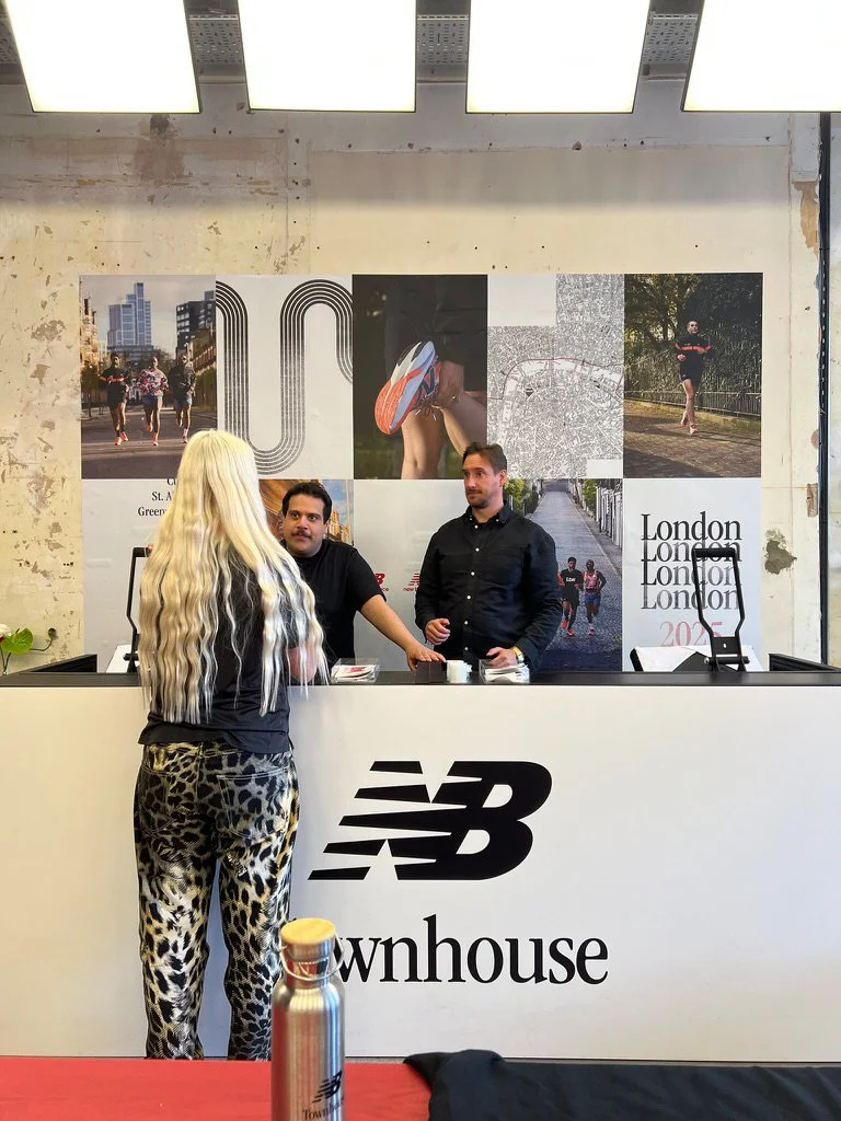A woman with long, wavy blond hair wearing leopard print pants and a black top is talking to two men behind a check-in counter. The counter has the New Balance logo and "townhouse" text. The background features large posters of running scenes in London and images of people jogging or standing outdoors.