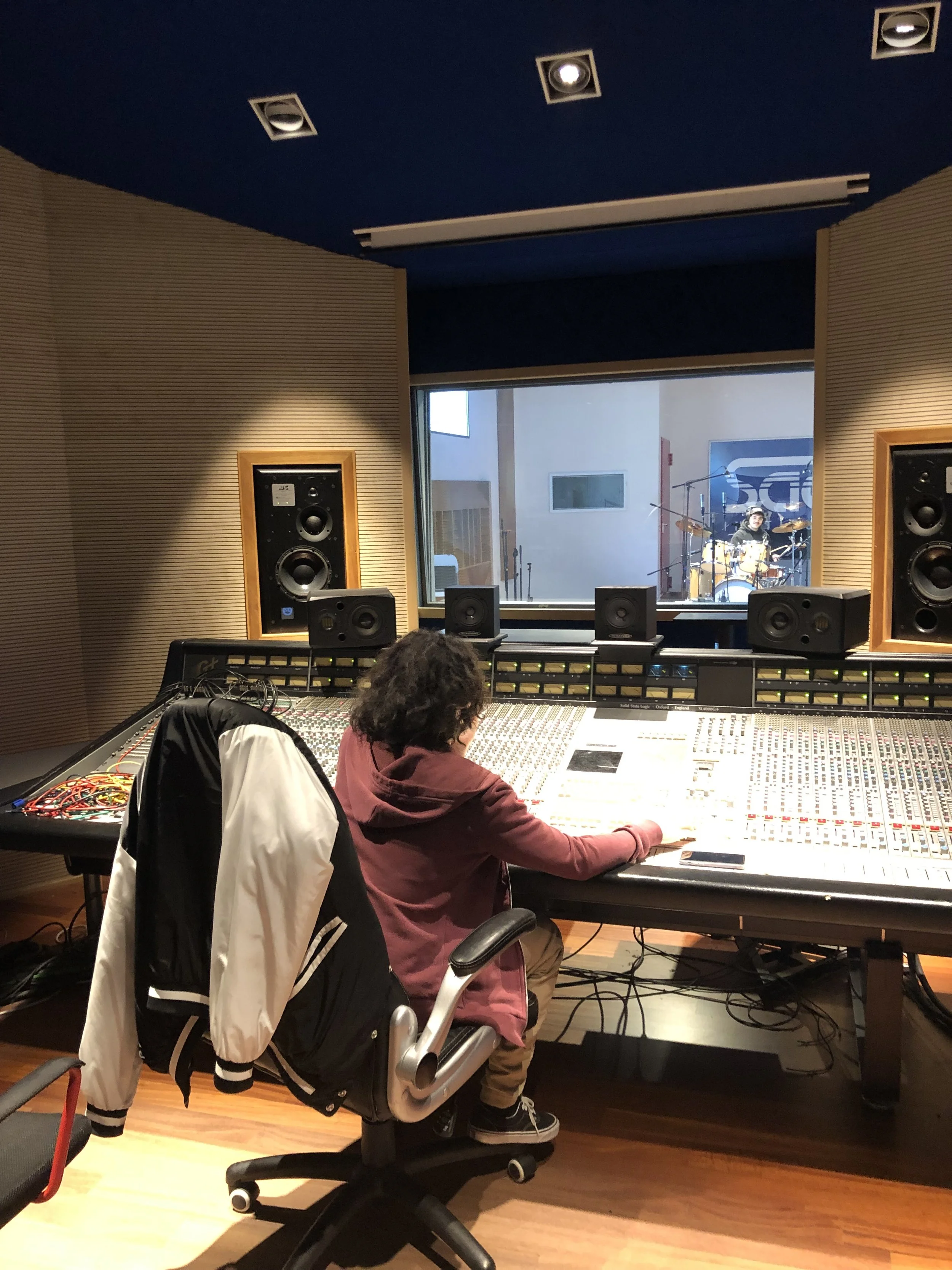Stanza di registrazione con una persona seduta davanti a un grande mixer audio, guardando attraverso una finestra verso un batterista che suona in un'altra stanza.
