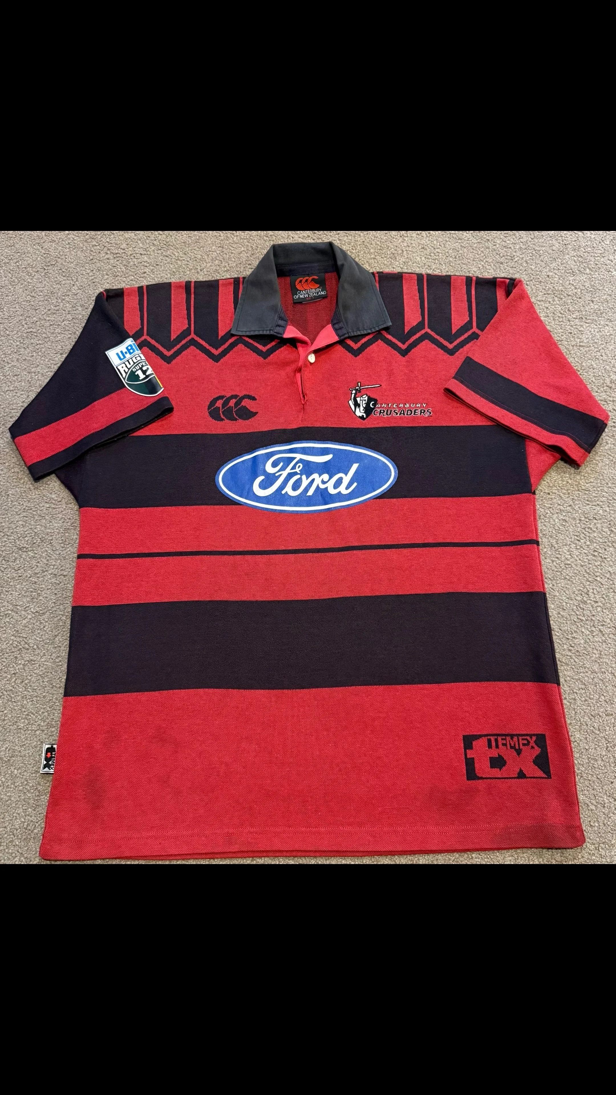 Red and black Canterbury Crusaders rugby jersey with sponsor logos and team branding laid flat on beige carpet.