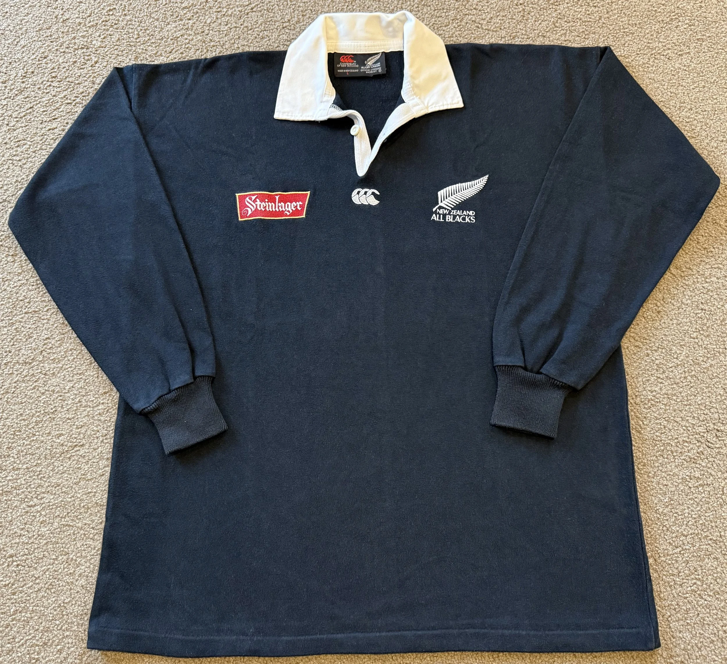 A black rugby shirt with a white collar, featuring logos for Steinglager, Canterbury, and New Zealand All Blacks on the front, laid flat on beige carpet.