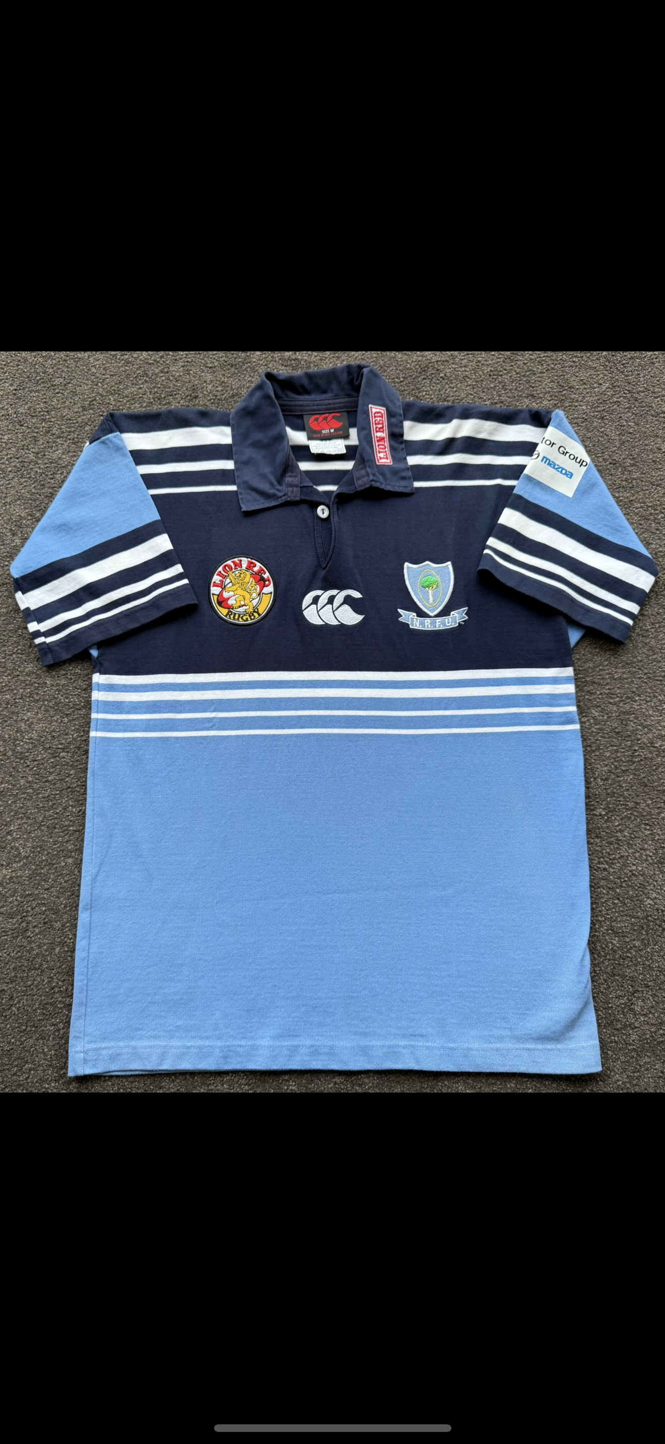 Blue rugby shirt with navy and white stripes and patches on the chest and sleeve, laid flat on a brown surface.