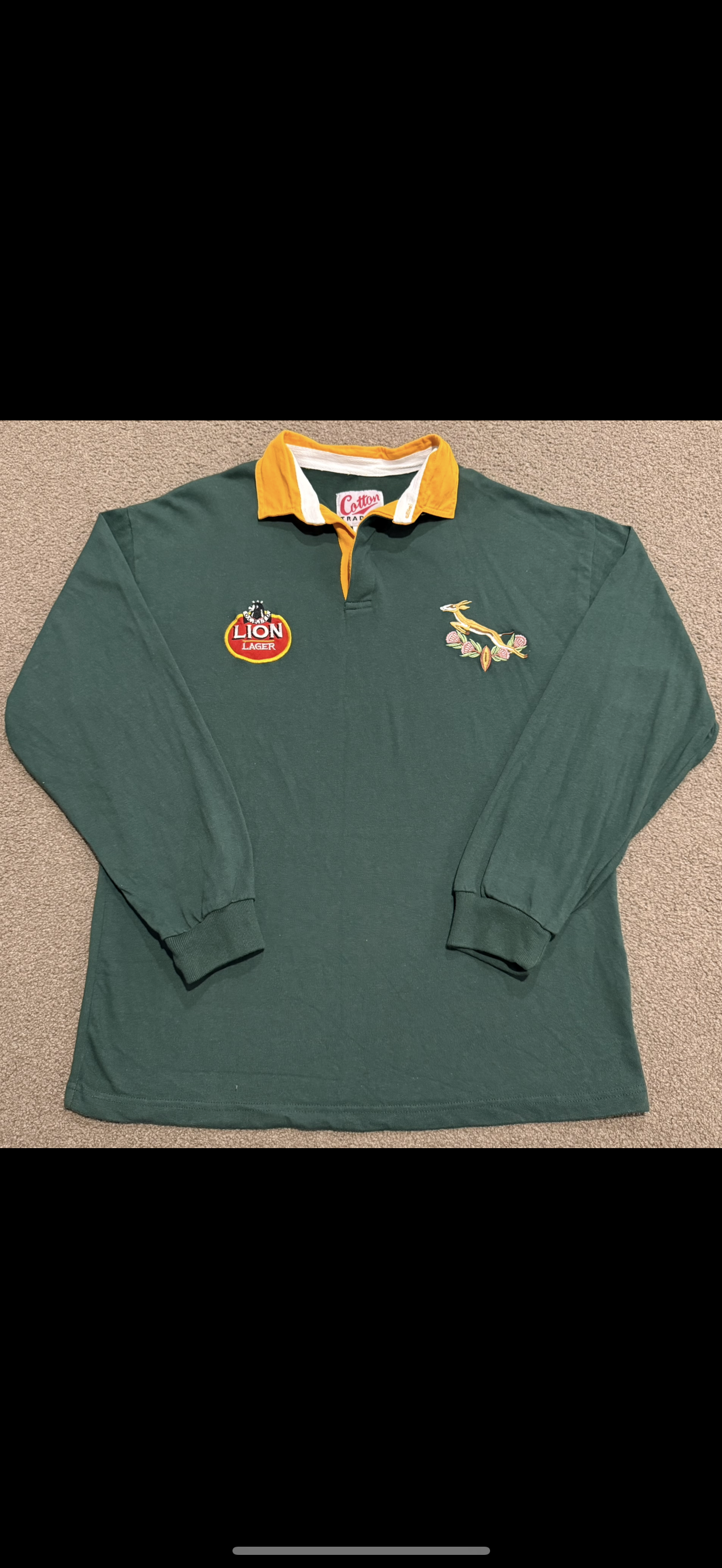 Green long-sleeve rugby shirt with yellow collar accents, embroidered Lions logo on left chest, and Lion Lager patch on right chest, lying on beige carpet.