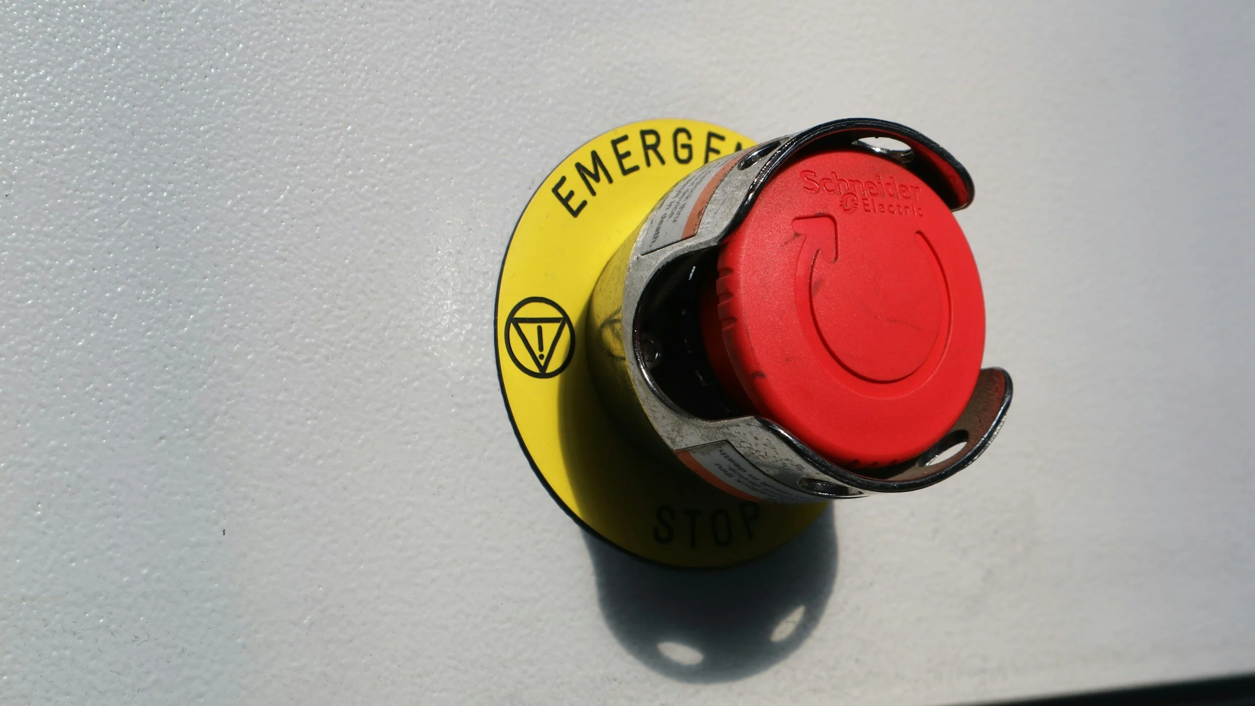 An emergency stop button with a red cap labeled 'Schneider Electric,' mounted on a white textured wall, with a yellow circular label underneath that reads 'EMERGENCY STOP.'