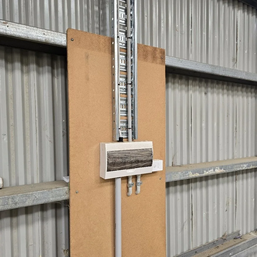 Electrical wiring and panel mounted on a corkboard behind a metal safety partition in an industrial setting.