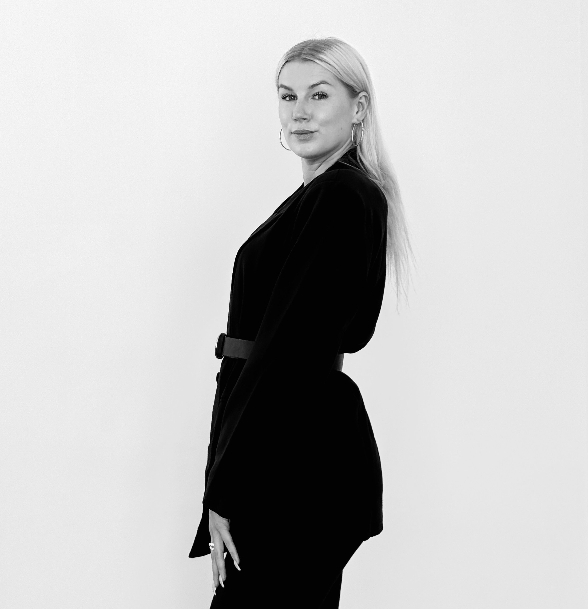 A black-and-white photo of a woman with long blonde hair wearing a black blazer and large hoop earrings, standing against a plain white background.