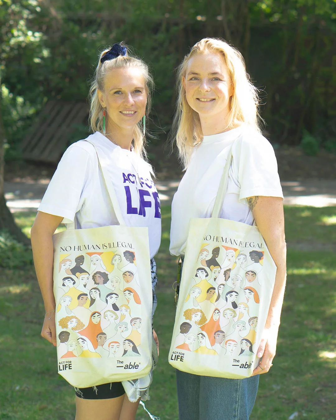 To kvinder står udendørs med ensjede totebags, der har tegn og teksten 'NO HUMAN IS ILLEGAL'.