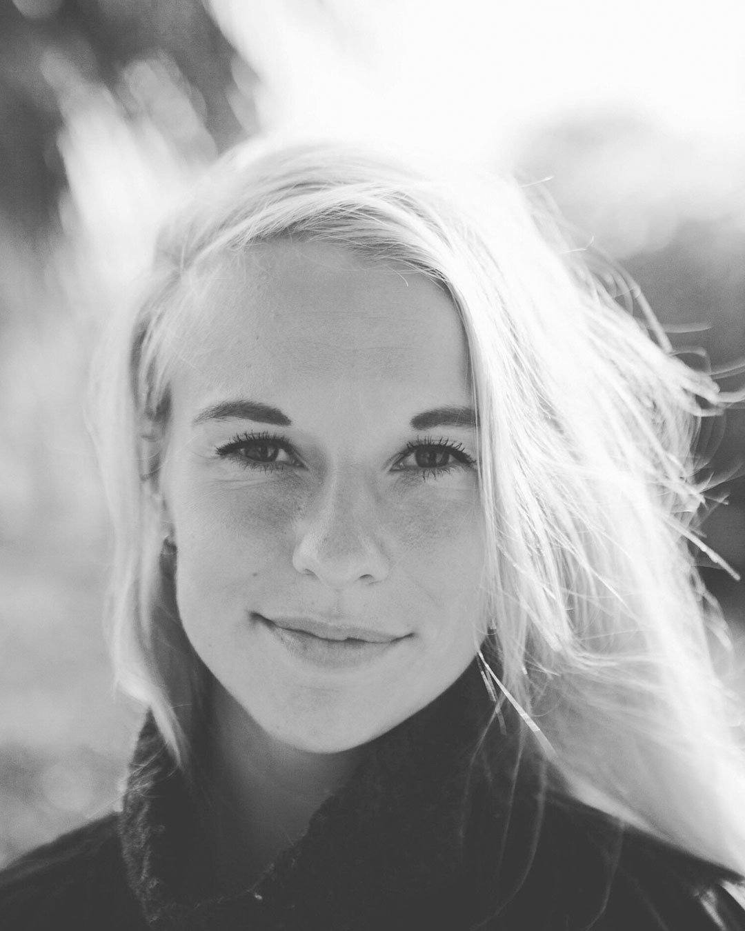 Sort-hvid portræt af en smilende ung kvinde med lyst hår, uden makeup, iført sort tøj, sollys, udkant i baggrunden