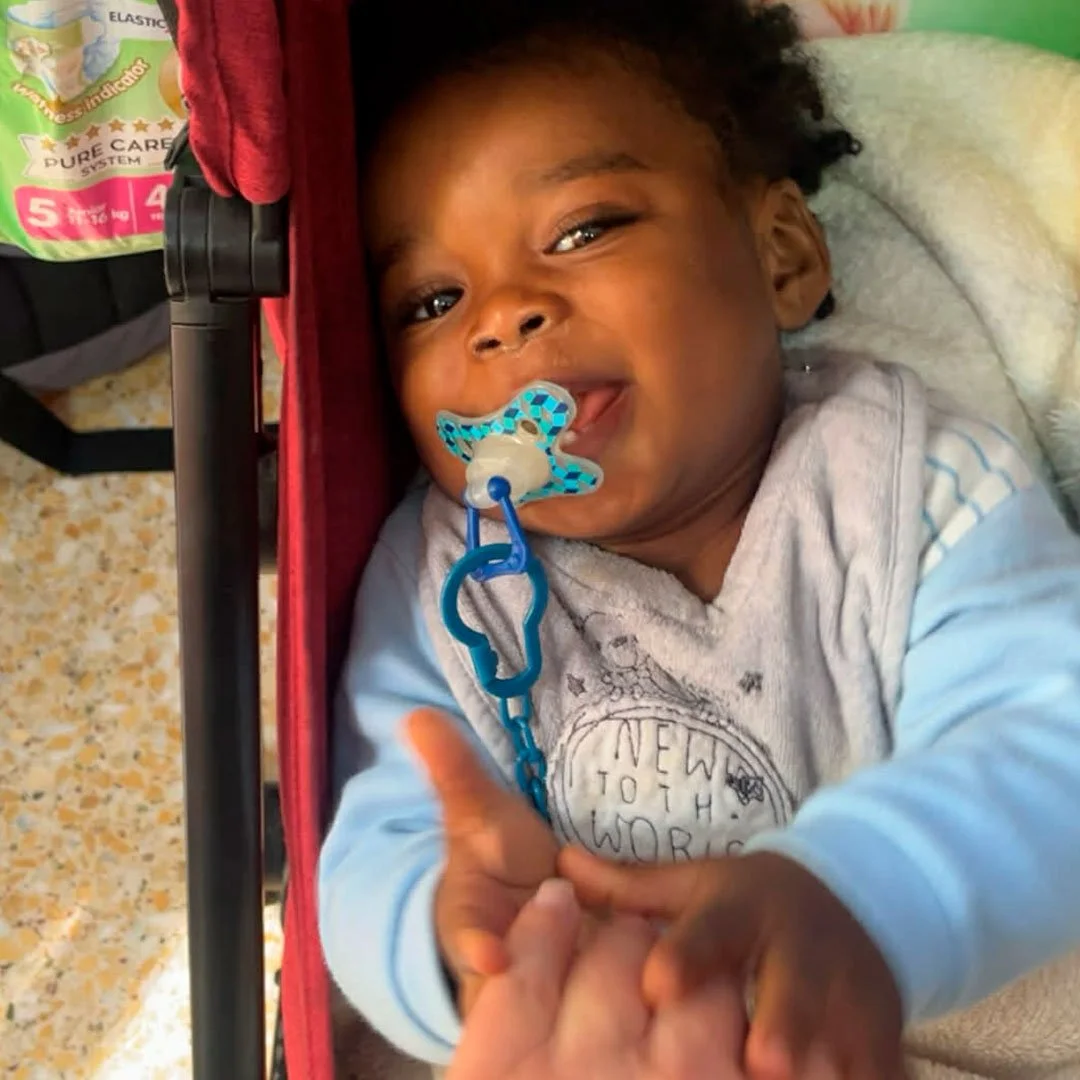 Smilende lille dreng med pacifier i munden, der giver håndtag til kameraet, liggende i barnevogn, iført gråt og blå tøj.