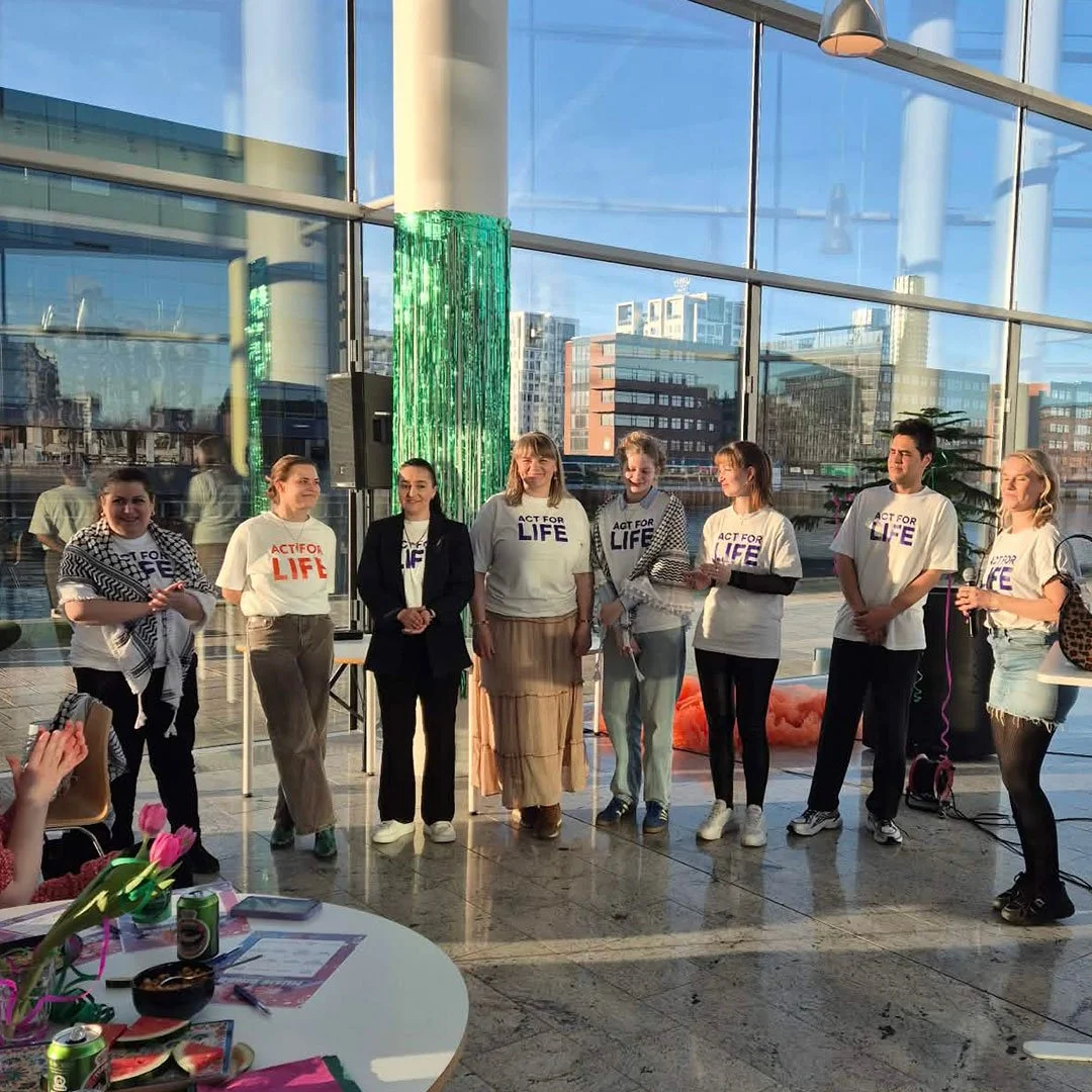 En gruppe mennesker står side om side indendørs foran store vinduer i et moderne byarkitekturmiljø. De er klædt i t-shirts med teksten 'ACT FOR LIFE' og ser ud til at deltage i et arrangement. Der er en bord med blomster, drinks og papirer i forgrunden.