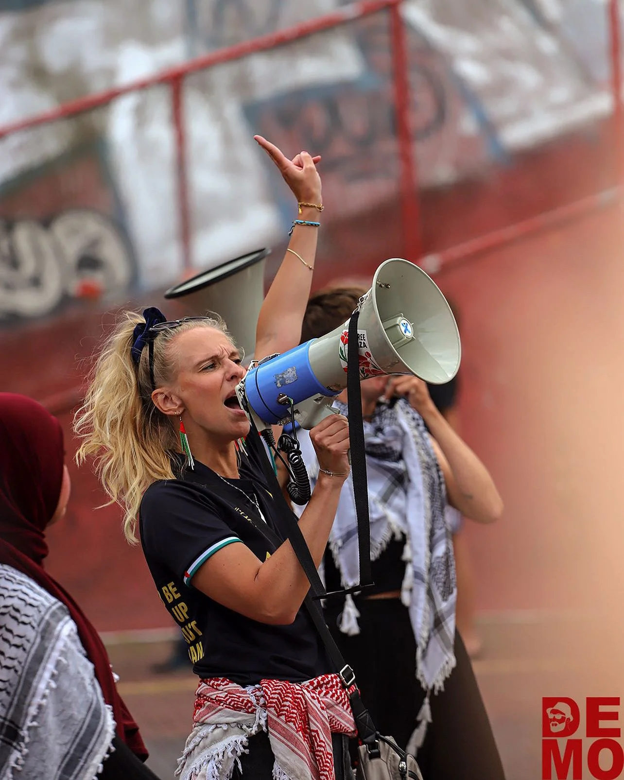 En kvinde med blond hår og farvestrålende øreringe holder en megafon og råber, omgivet af andre mennesker. Baggrunden viser et sportsanlæg med graffiti.