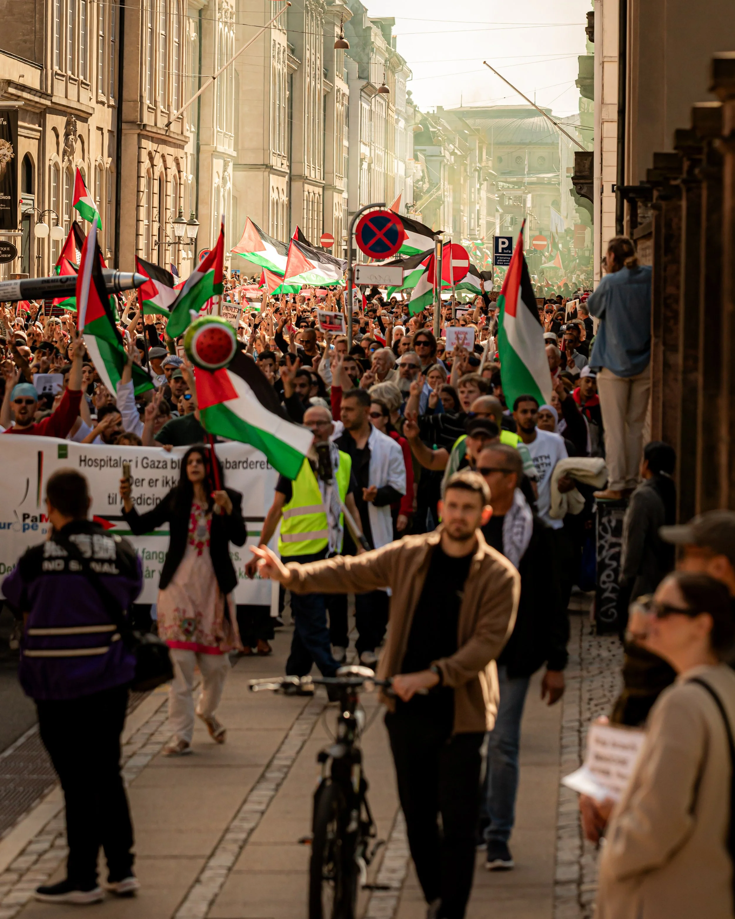 En stor gruppe mennesker protesterer på en gade, mange holder palestinske flag og skilte. Der er bygninger med arkitektur fra en europæisk by, og flere tager billeder eller ser på demonstrationen.