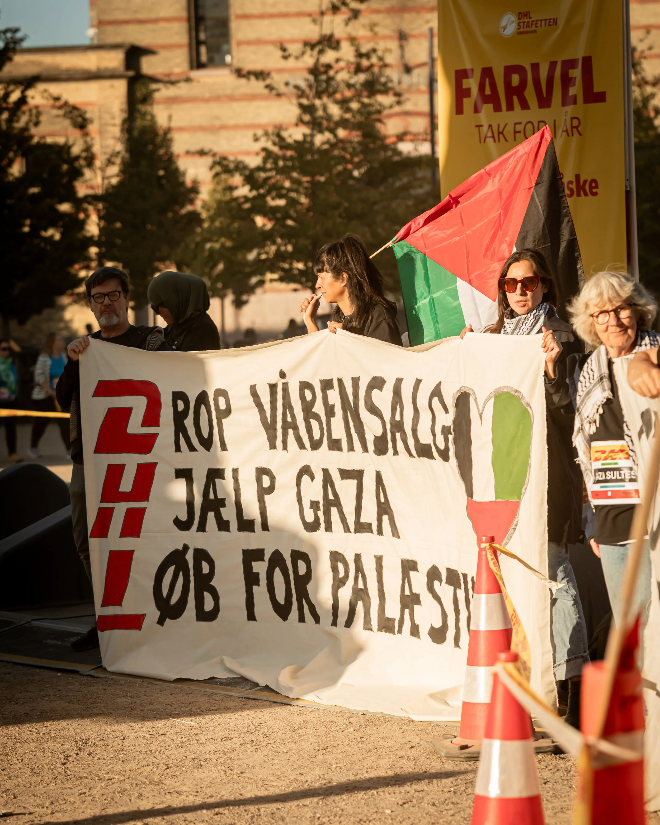 Protest foran med folk, store bannere og flag, der viser modstand mod forarbejdning af våben og atomvåben efter krigen i Gaza, med en bymur i baggrunden.