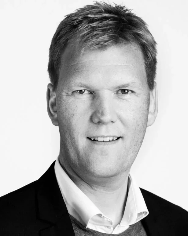 Sort-hvid portræt af en ung mand med kort, lyst hår iført hvid skjorte og sort jakke, smilende.
