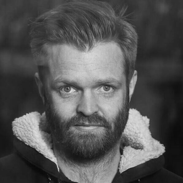 Sort-hvid portrætfotografi af en mand med lyst, kort hår, skæg og blå øjne, iført en hættetrøje.