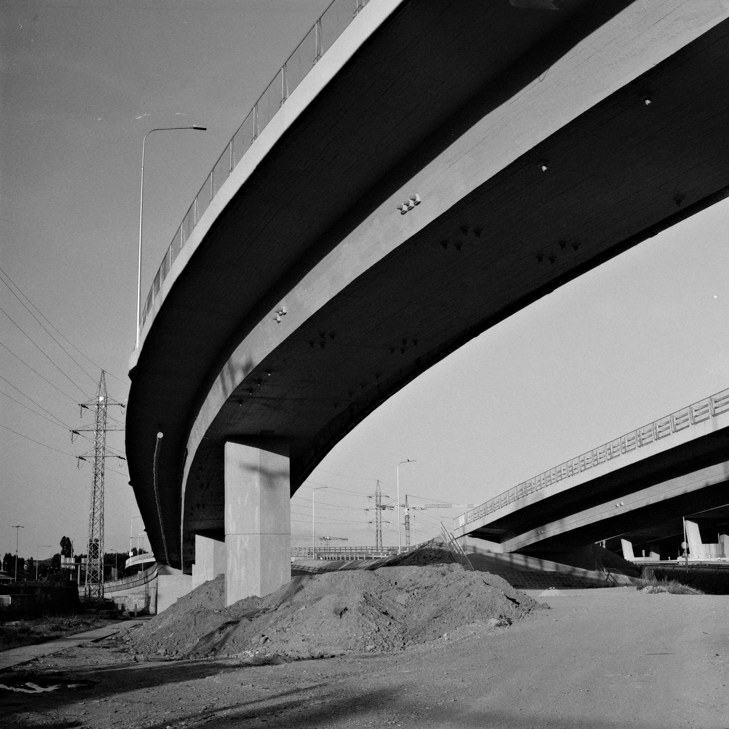 Vista di un viadotto in costruzione con pile di terra sottostanti e linee elettriche sullo sfondo.