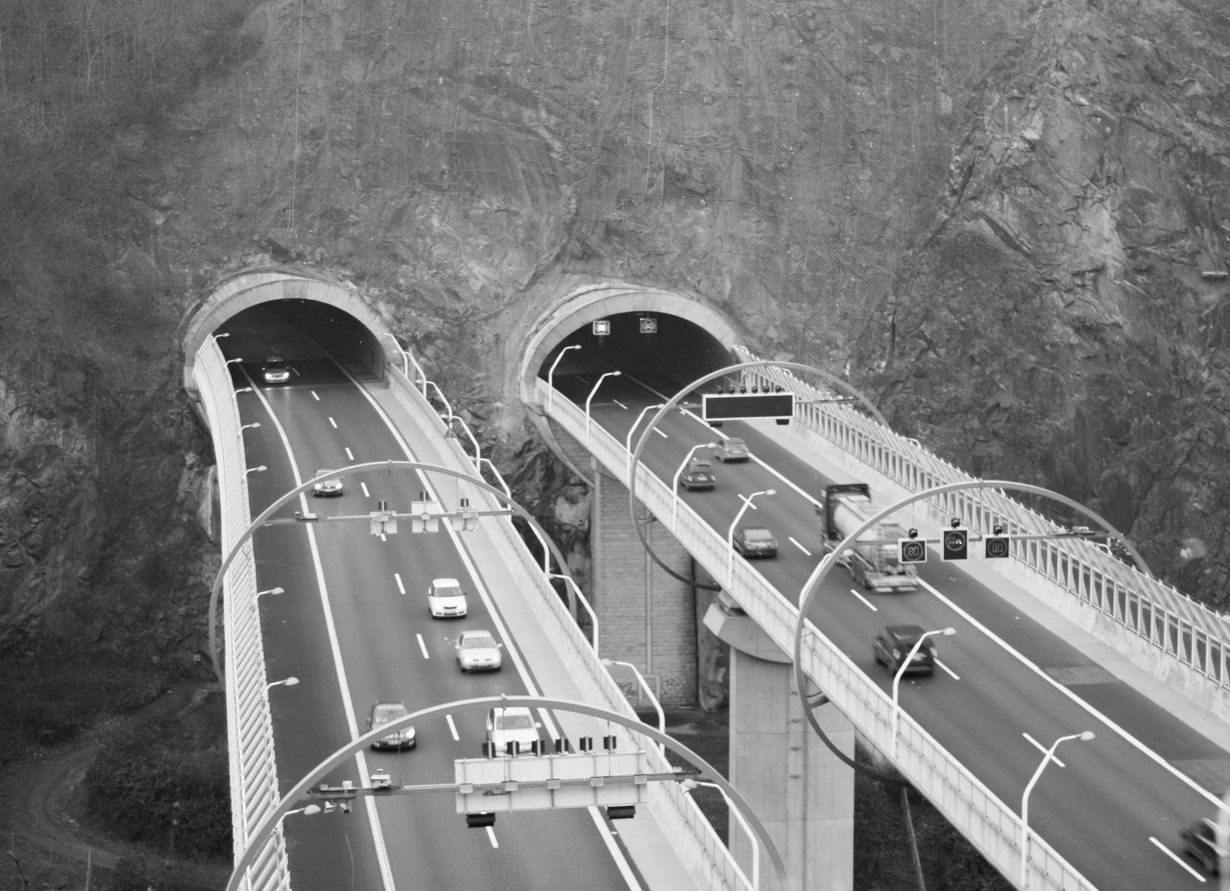 Immagine di una strada in montagna con due tunnel e diversi veicoli in movimento, in bianco e nero.
