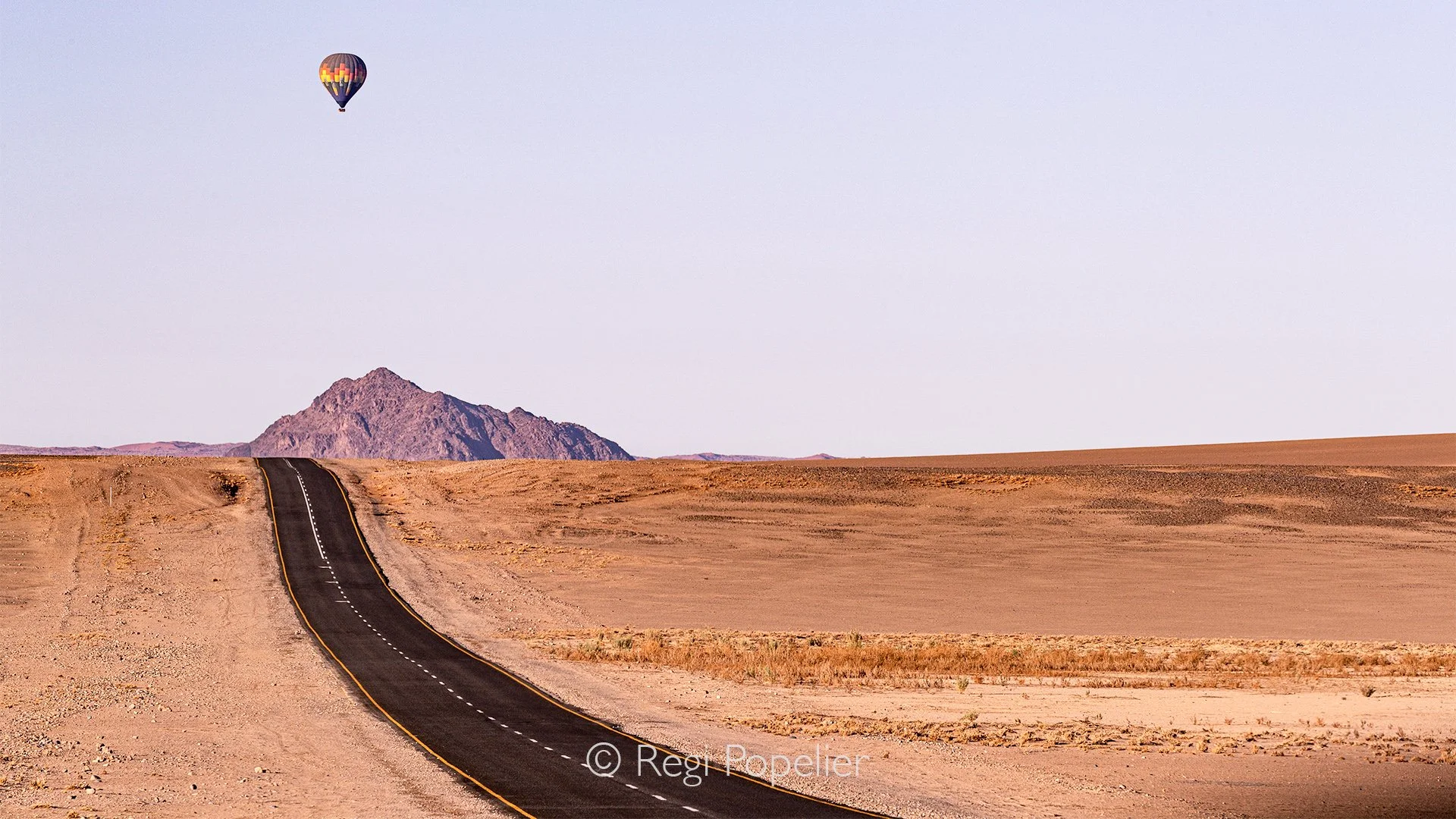 Namibia063 - A study in landscape contrast. The sleek, dark texture of the asphalt road provides a perfect visual anchor for the photographer. It highlights the vast scale of the hyper arid landscape, showing how modern engineering carves a path thro