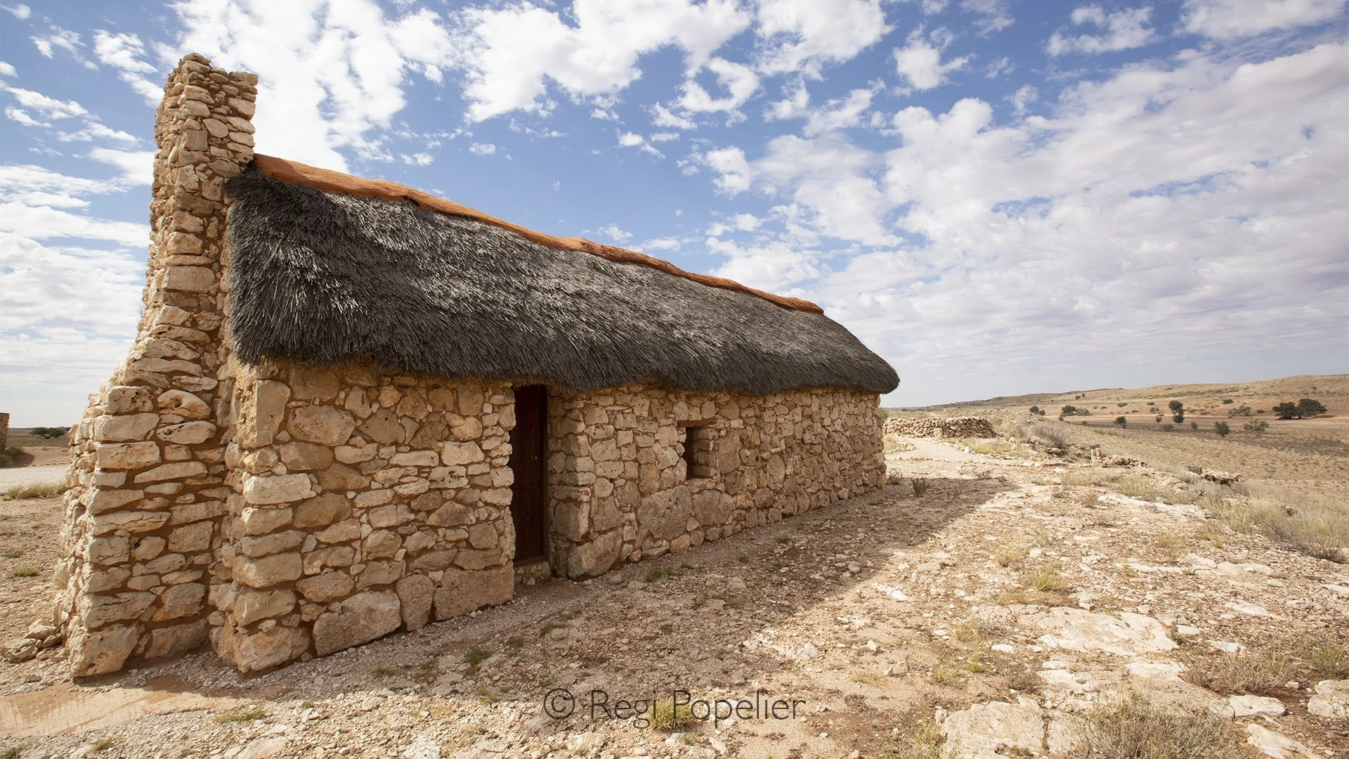 South Africa007 -  Amid the endless red sands of the Transfrontier Kalahari stands a humble stone house, weathered by decades of relentless sun, wind, and silence. Surrounded by sparse vegetation and an ocean of open wilderness, it seems almost forgo