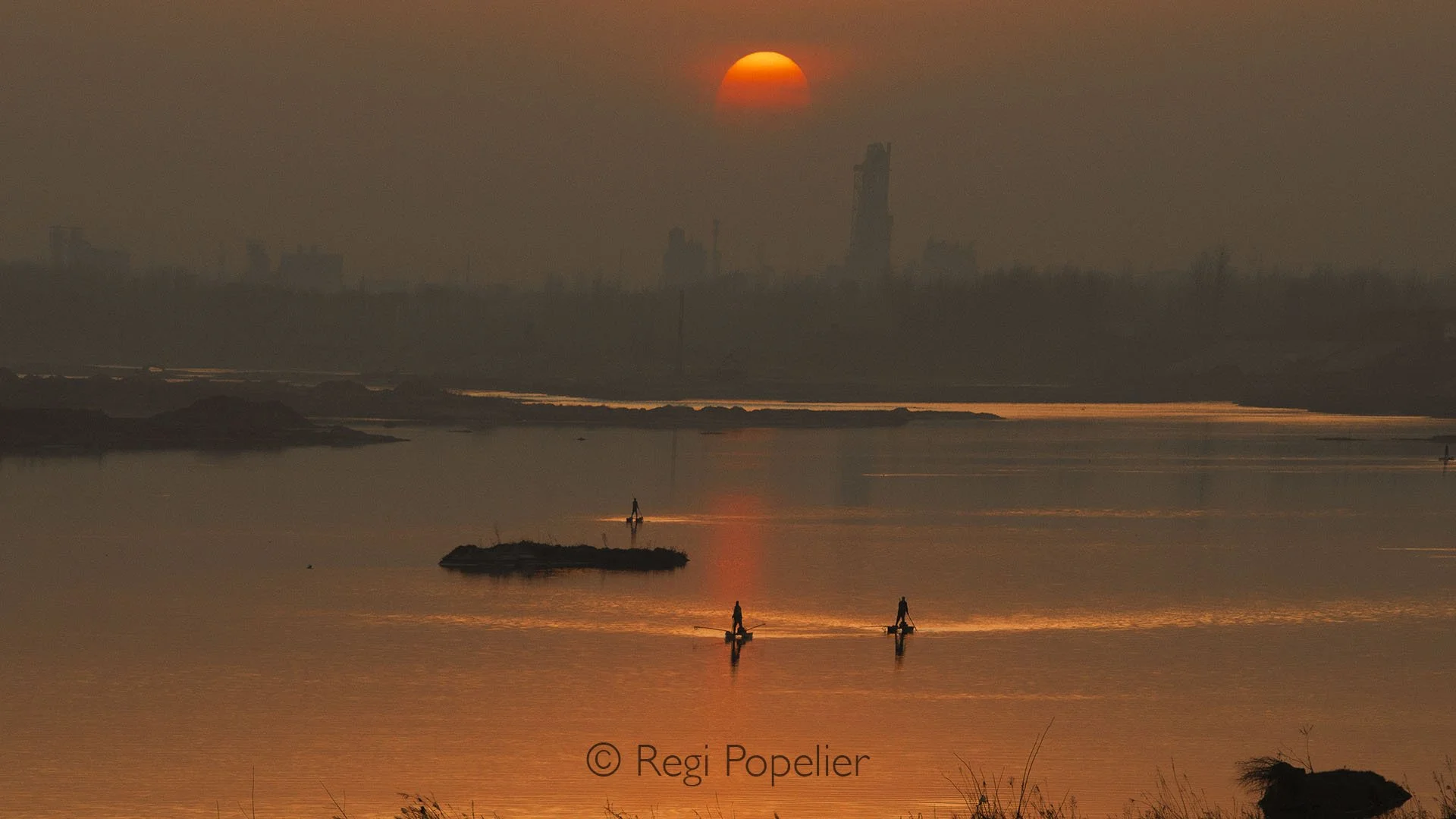 CHIN003-Fishermen make their way home after a long day on the river, their silhouettes drifting across the glowing water as the sun sets in warm hues behind them.