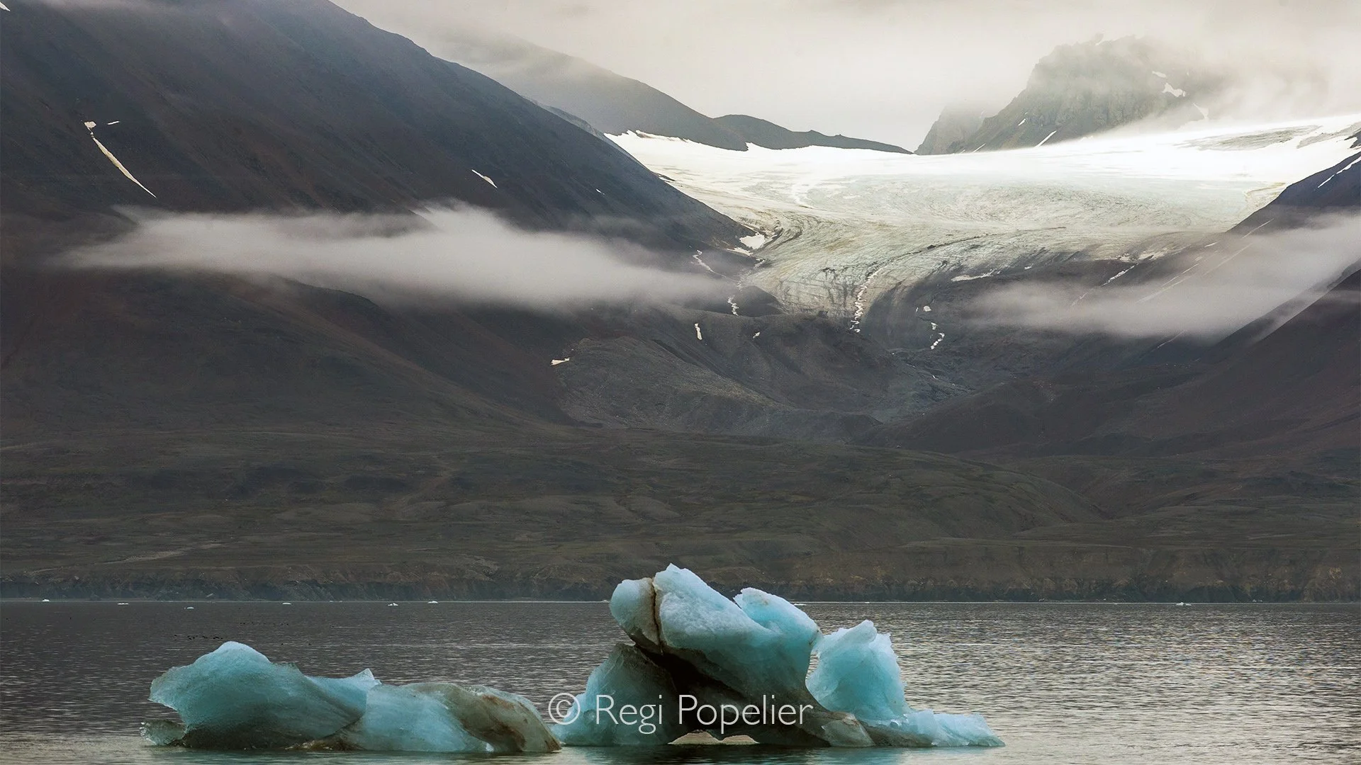 NPOLE016 - A lone iceberg drifts silently through the fjord beneath towering mountains.