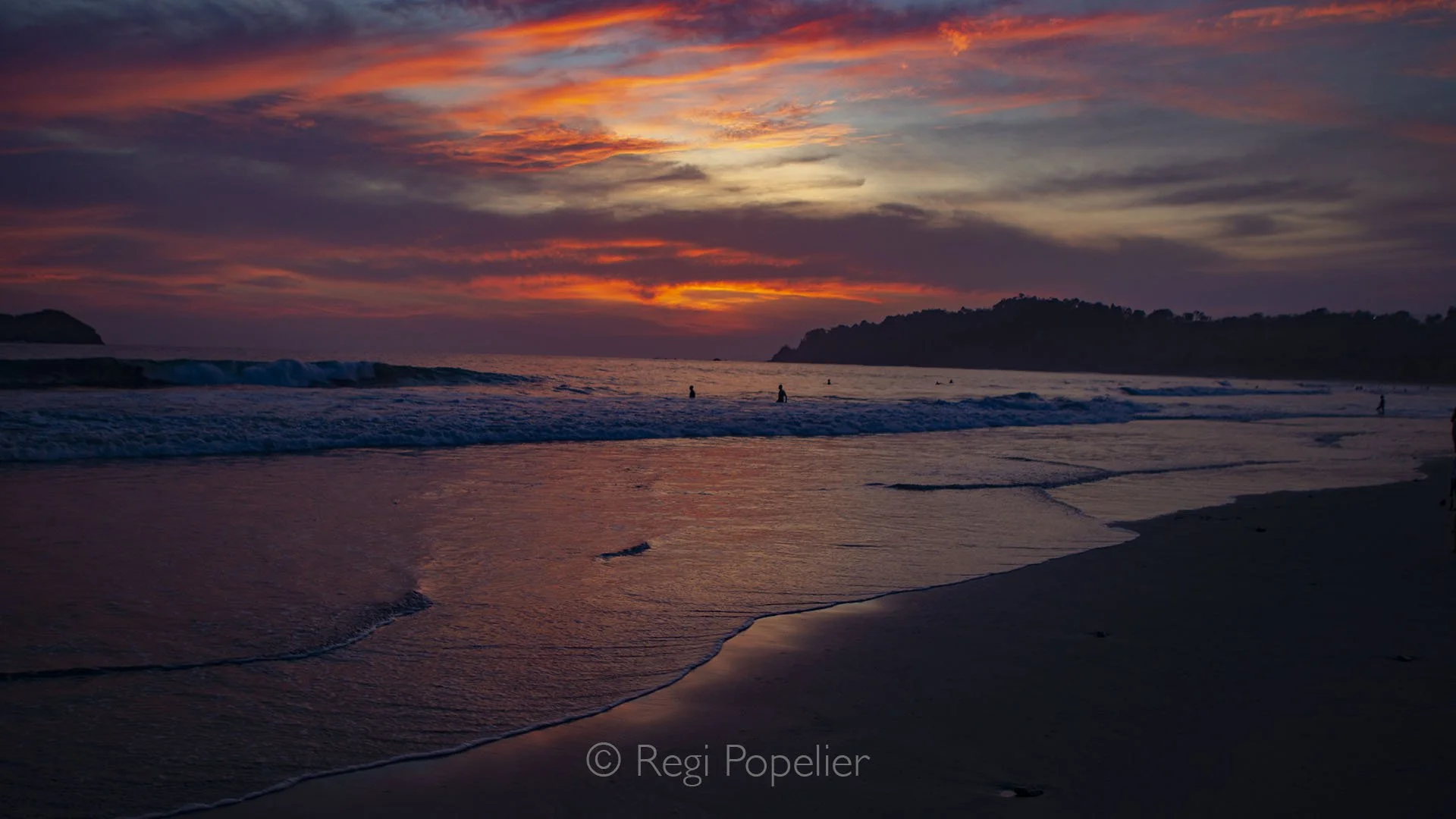 CR021 - People stroll along the beach, their silhouettes dark against the fiery red and orange hues of the setting sun, creating a serene and dramatic coastal scene.