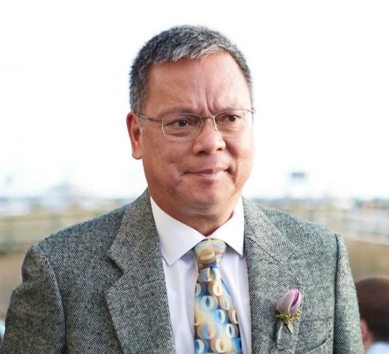 A man wearing glasses, a gray suit, a white shirt, and a colorful tie with a flower pinned to his lapel, standing outdoors.