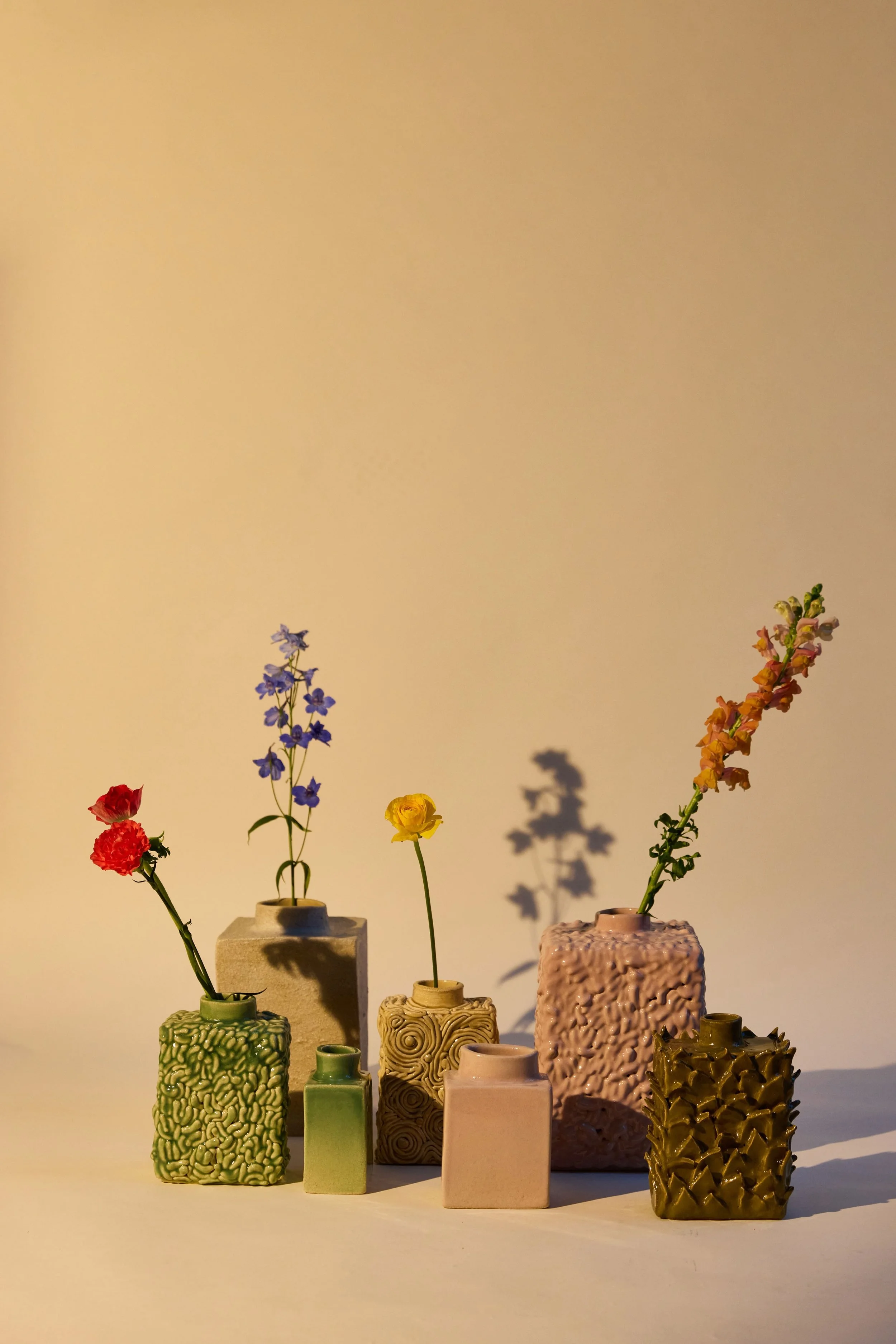 Vases en céramique de différentes tailles et formes, contenant des fleurs colorées, sont placés sur une surface lumineuse avec un fond neutre. Les vases ont des textures variées, allant de rugueuses à lisses, et chaque vase contient une unique fleur ou un bouquet.