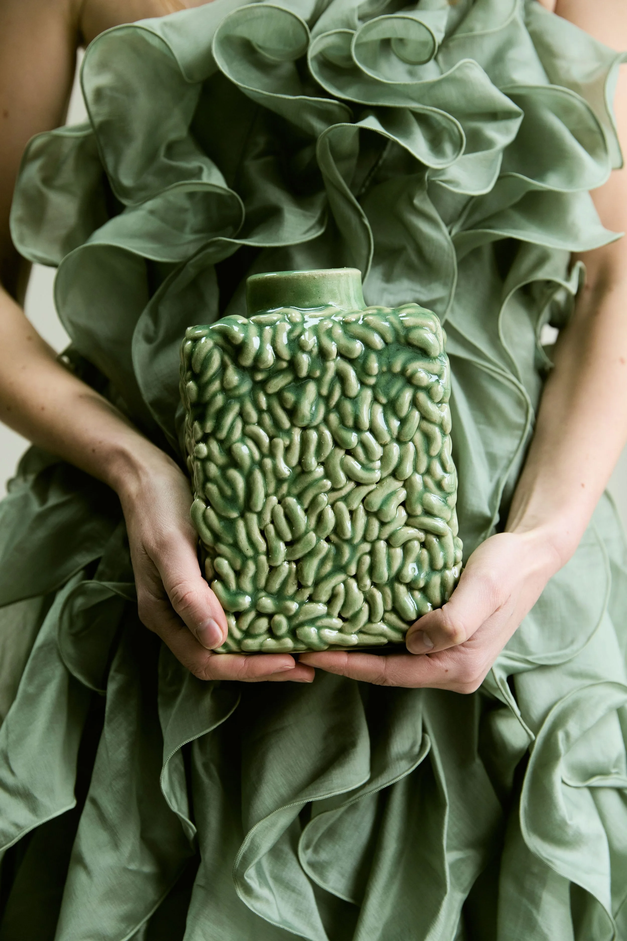 Personne portant une robe  verte avec des volants, tenant un vase vert brillant avec une texture en forme de cerveau.