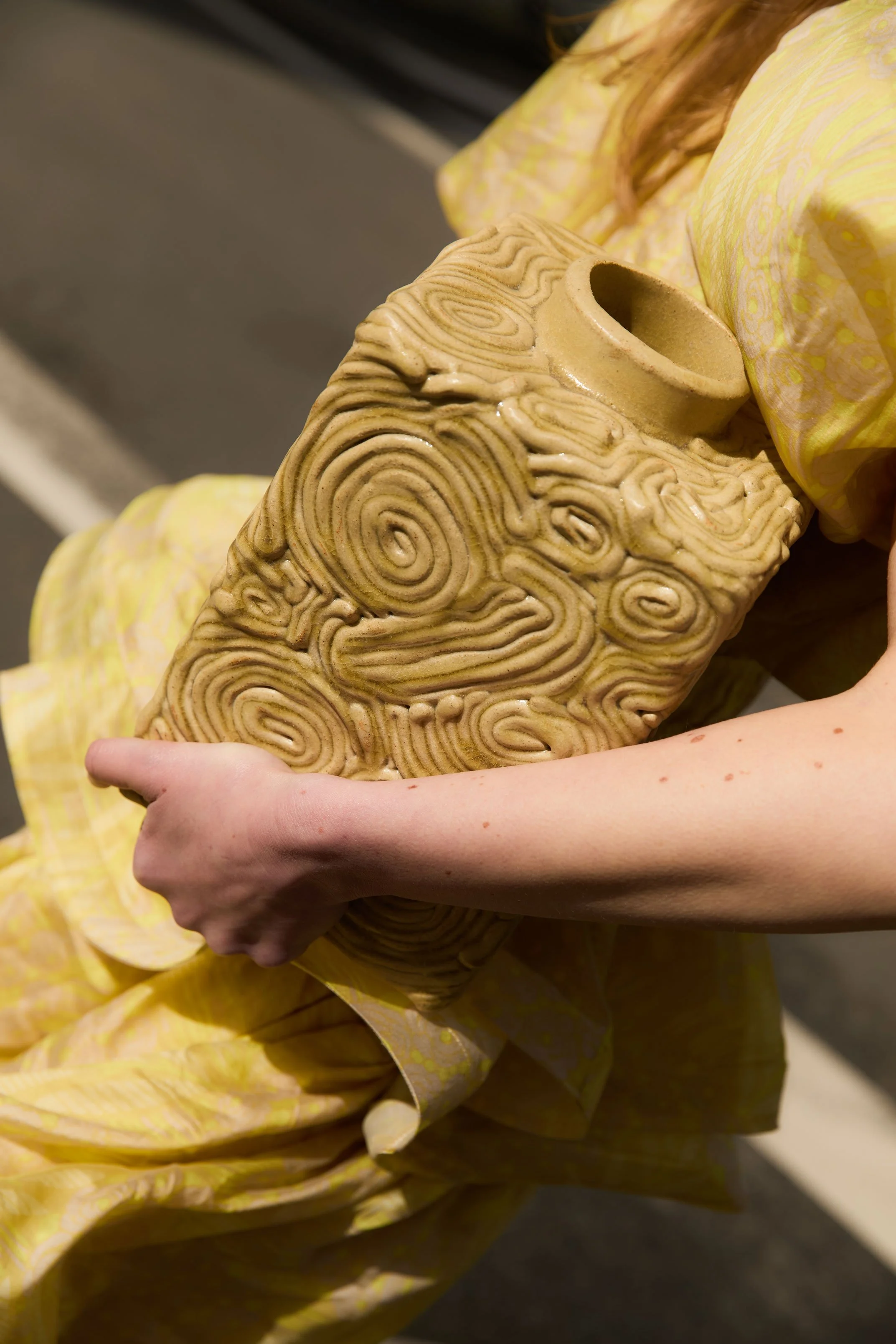 Une personne tenant une sculpture en céramique beige avec des motifs de spirales et de lignes ondulées, tout en étant vêtue d'une robe jaune à motifs.